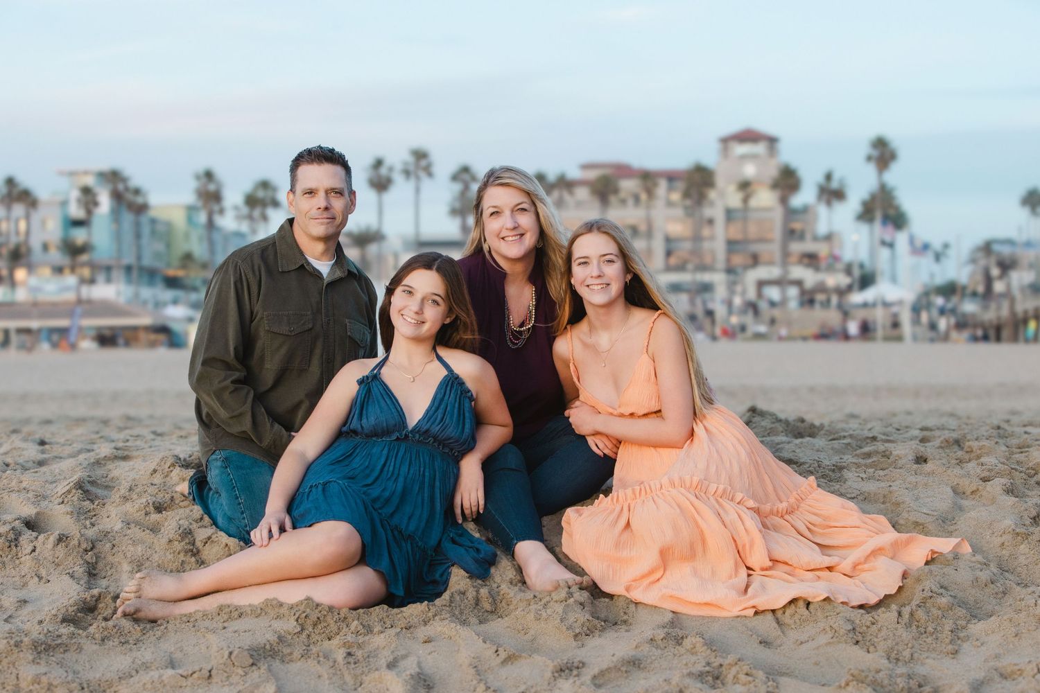 Family of four smiling together on a sandy beach at sunset with palm trees and colorful buildings in the background.