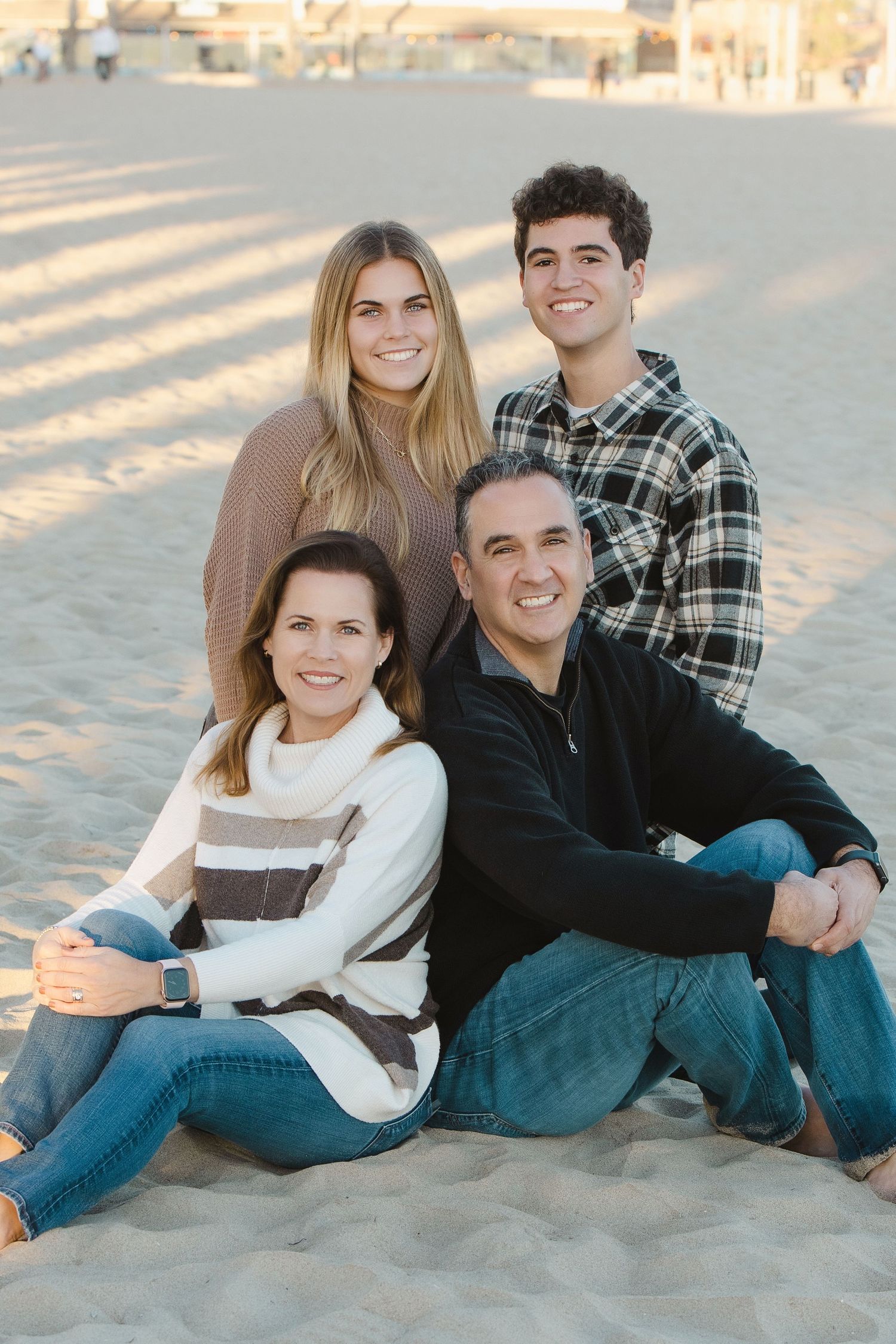 Happy family of four smiling together on a sandy beach, wearing casual sweaters and jeans in warm afternoon light.