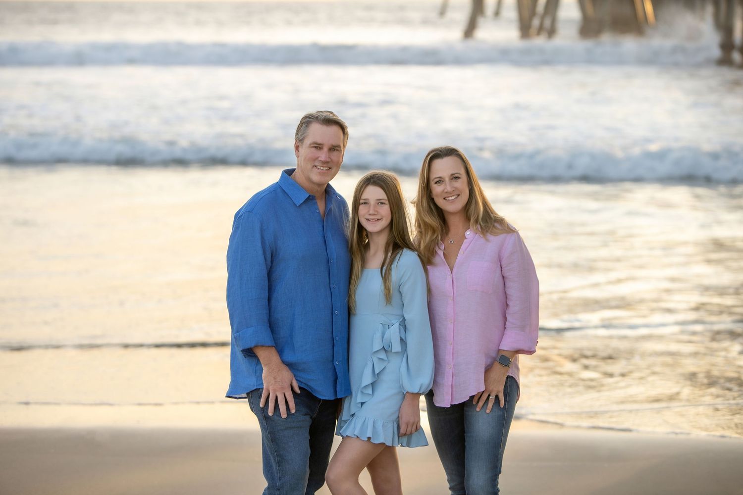 Family of three smiling on a sandy beach at golden hour with ocean waves in the background.