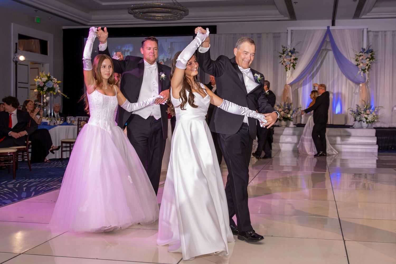Bride and groom dancing with wedding party on elegant ballroom dance floor during formal wedding reception.
