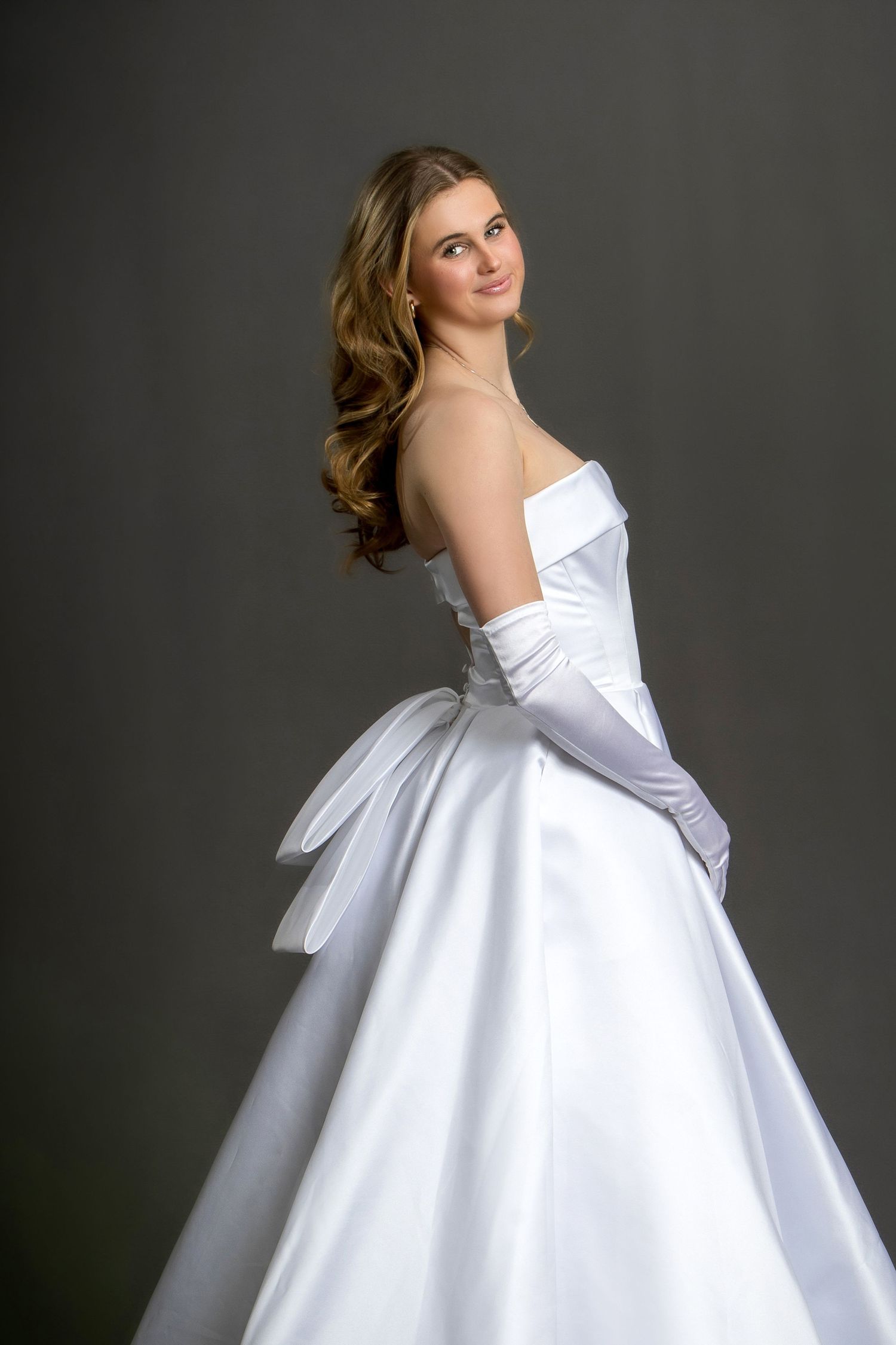 Elegant bride in a white strapless satin ball gown with long gloves and bow detail, posing gracefully in studio.