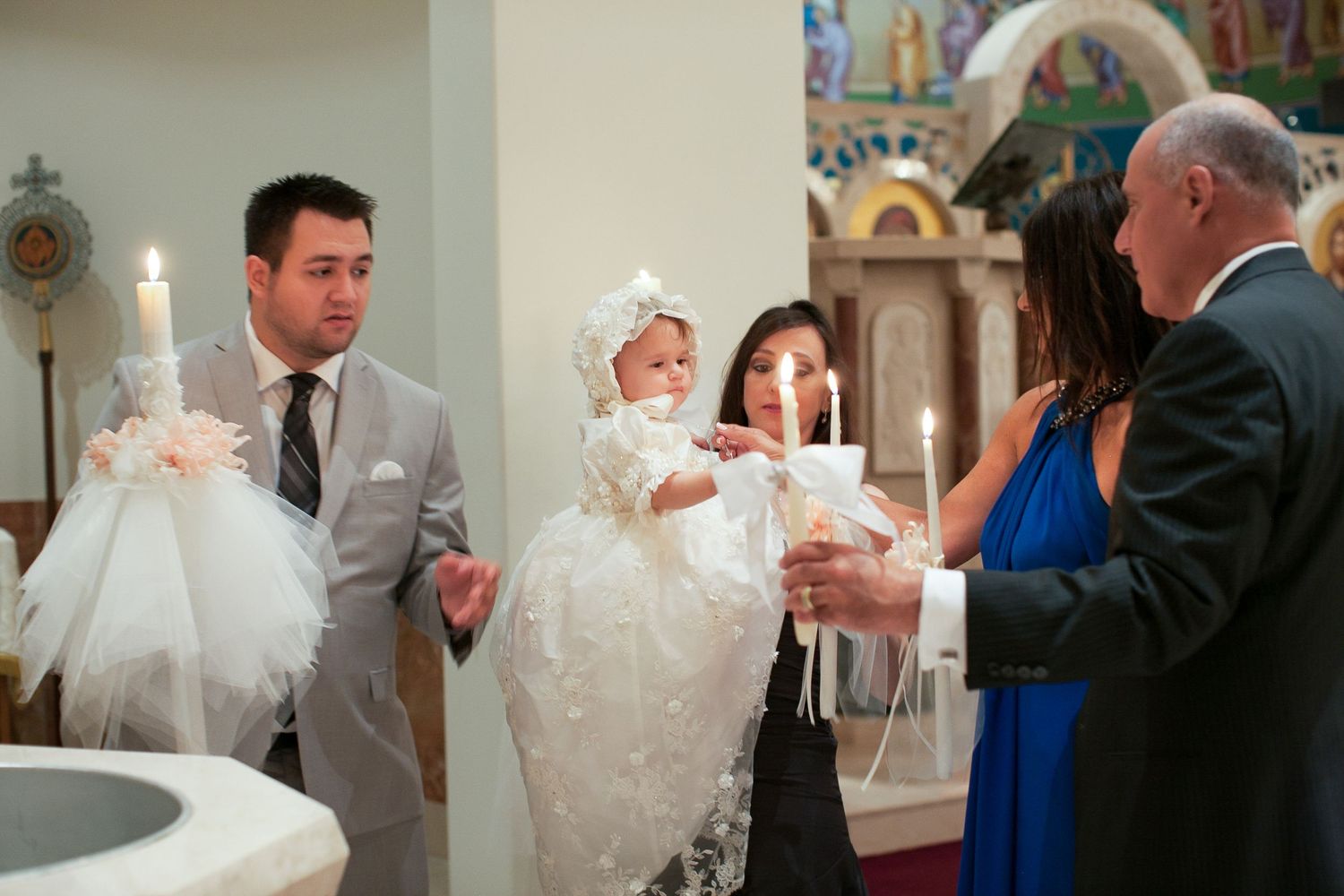 Baby in white christening gown held during baptism ceremony with family holding candles in church.