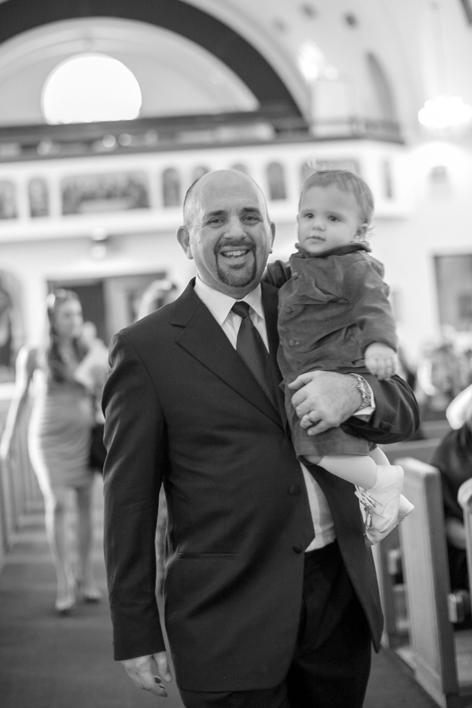 Man in suit holding a baby smiles while walking down a church aisle in a black and white photo.