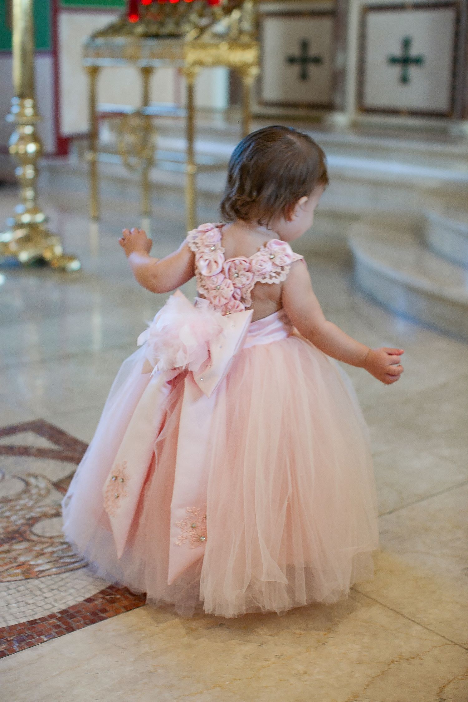 Toddler girl in pink tulle ball gown with floral back detail standing in elegant church interior.