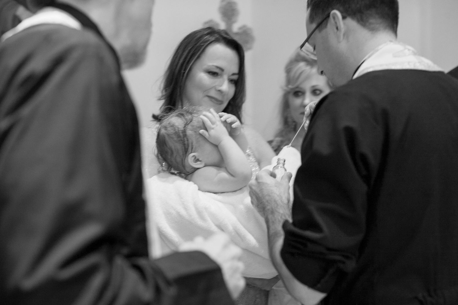 Black and white photo of a woman holding a baby during a baptism ceremony with a priest and guests nearby.