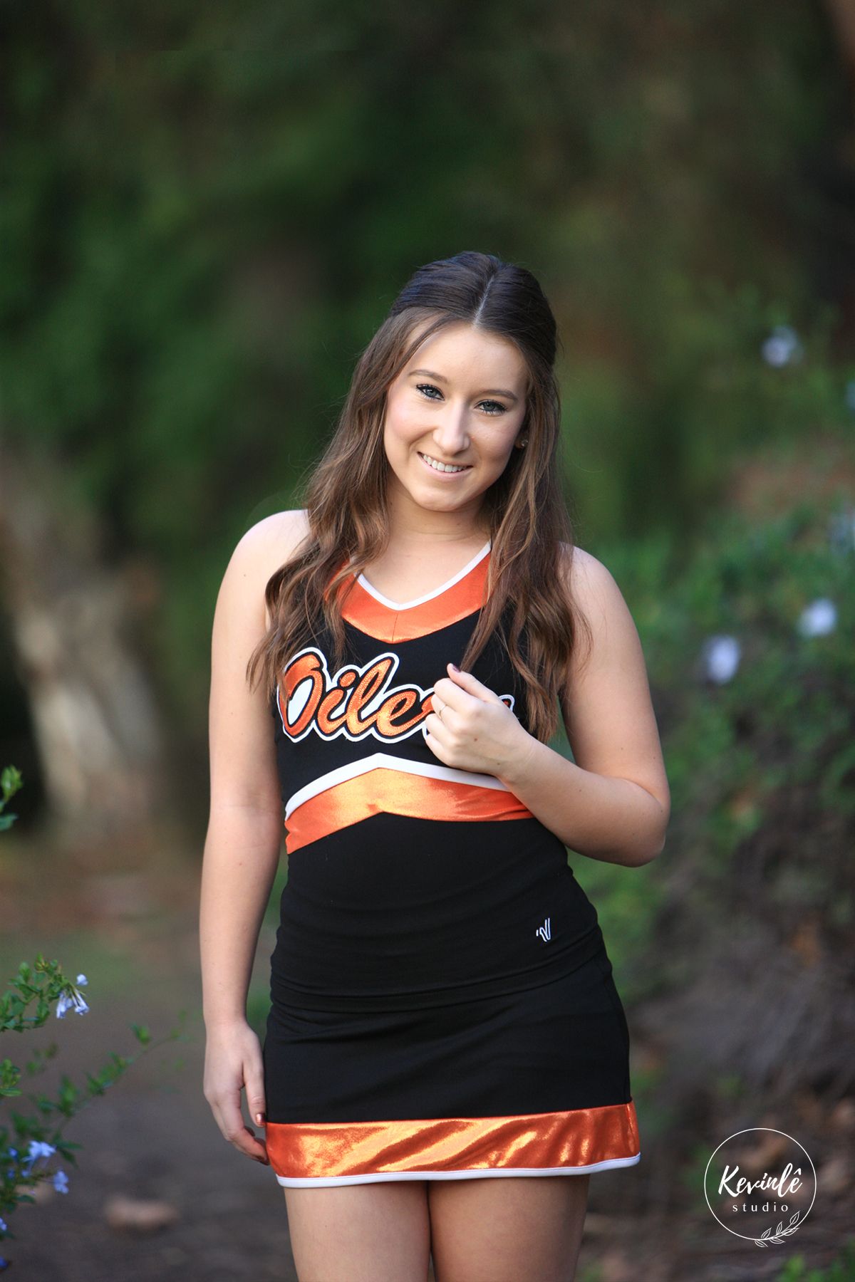 Young woman in black and orange Oilers cheerleader uniform smiling outdoors in a garden setting.