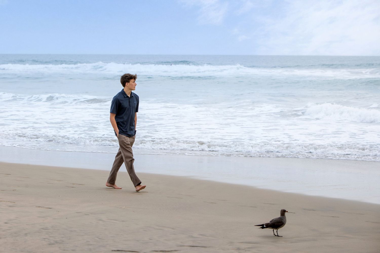 Man walking barefoot on sandy beach near ocean waves with a seagull nearby on a cloudy day.