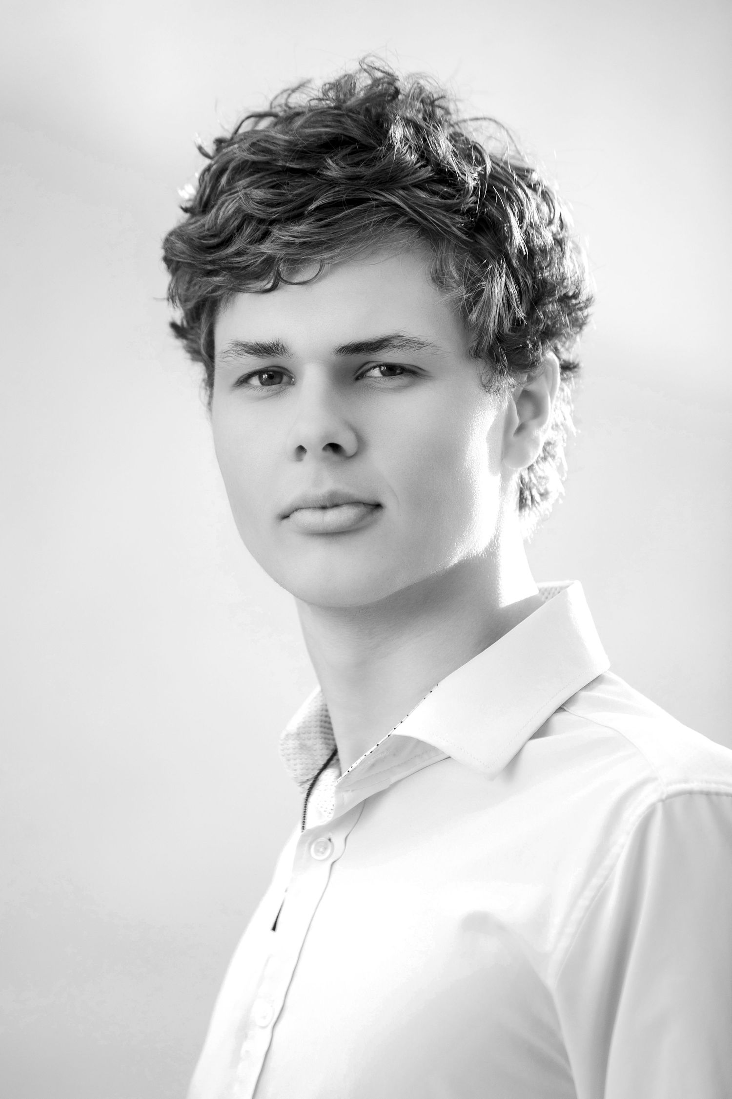 Black and white portrait of a young man with curly hair wearing a white dress shirt, looking directly at the camera.