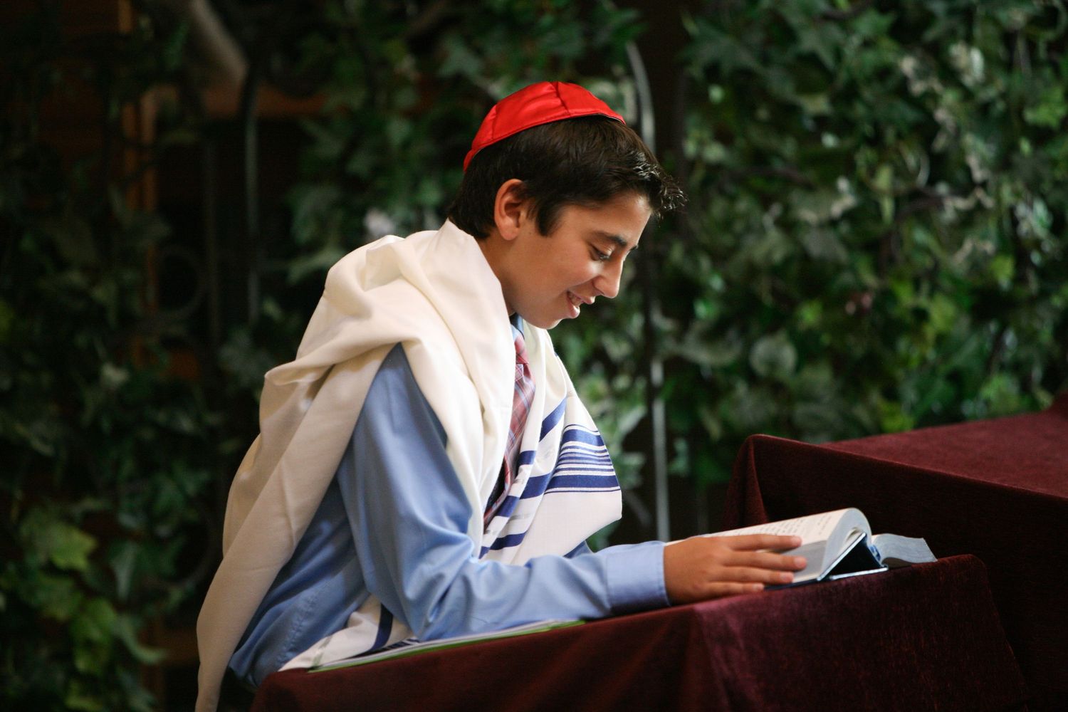 Young Jewish boy in red kippah and tallit reads from Torah during Bar Mitzvah ceremony outdoors.