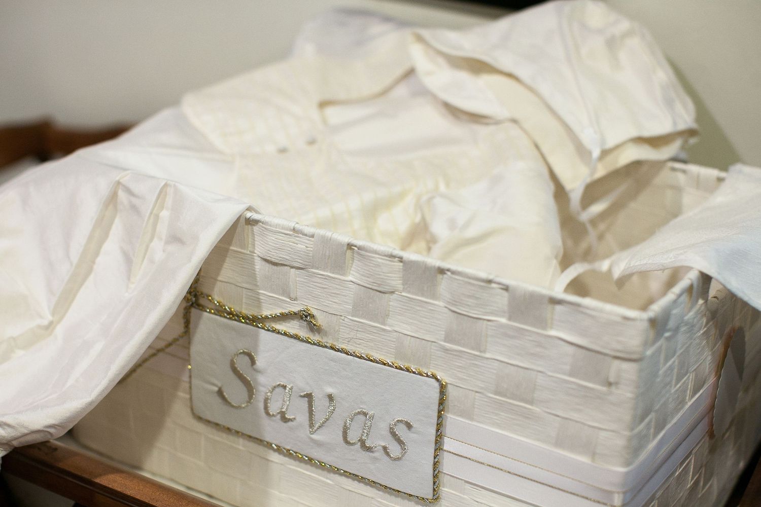 White wicker storage box labeled Savas filled with white fabric or linens on a wooden surface.