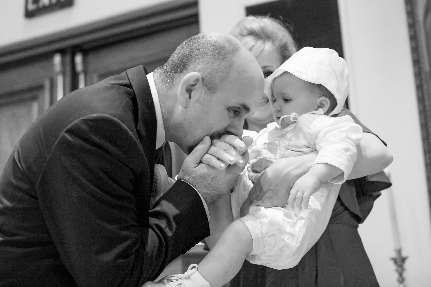 Black and white photo of a man in a suit kissing a baby's hand at a christening ceremony.