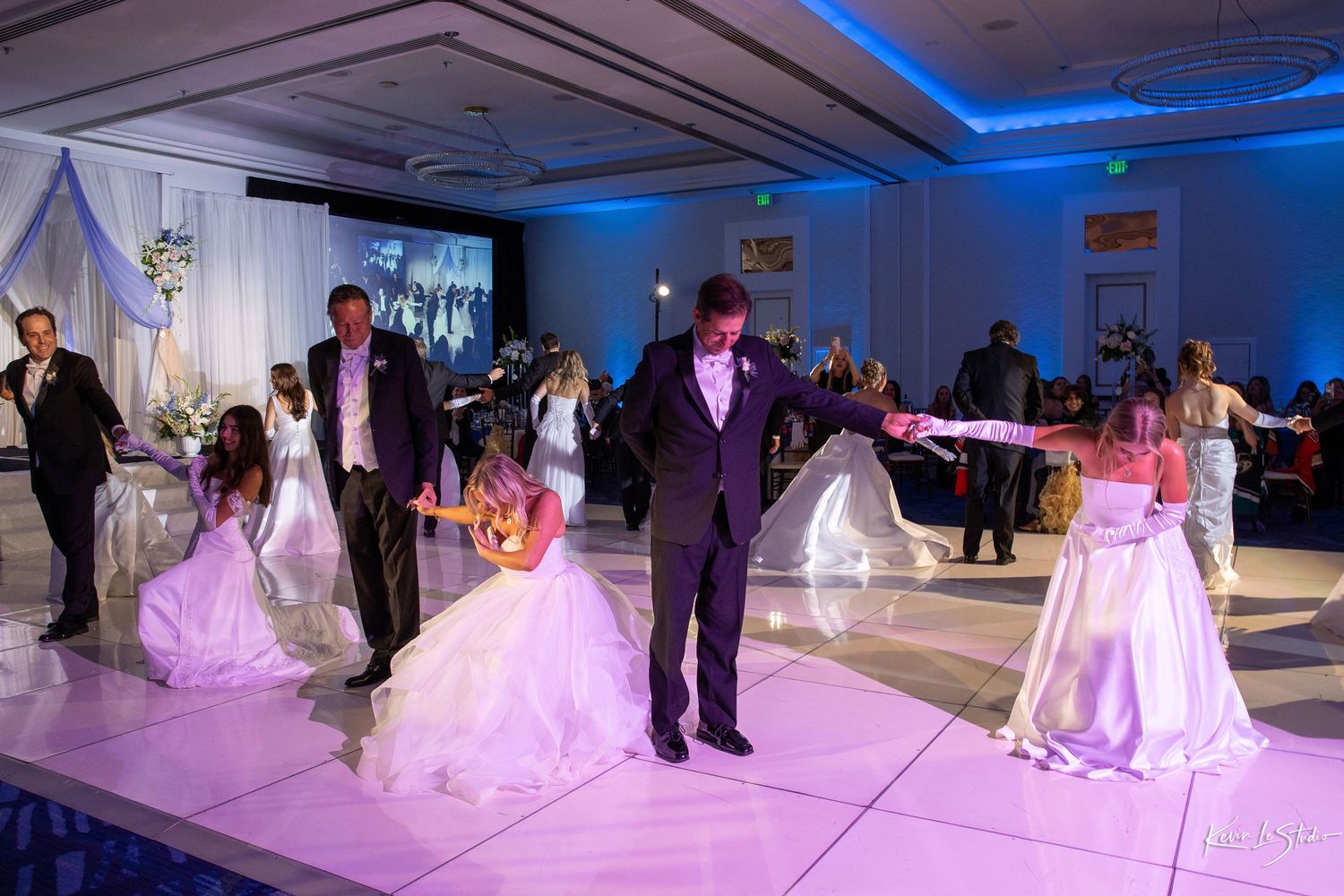 Elegant wedding reception dance floor with bride and groom dancing alongside guests in formal attire under blue ambient lighting.