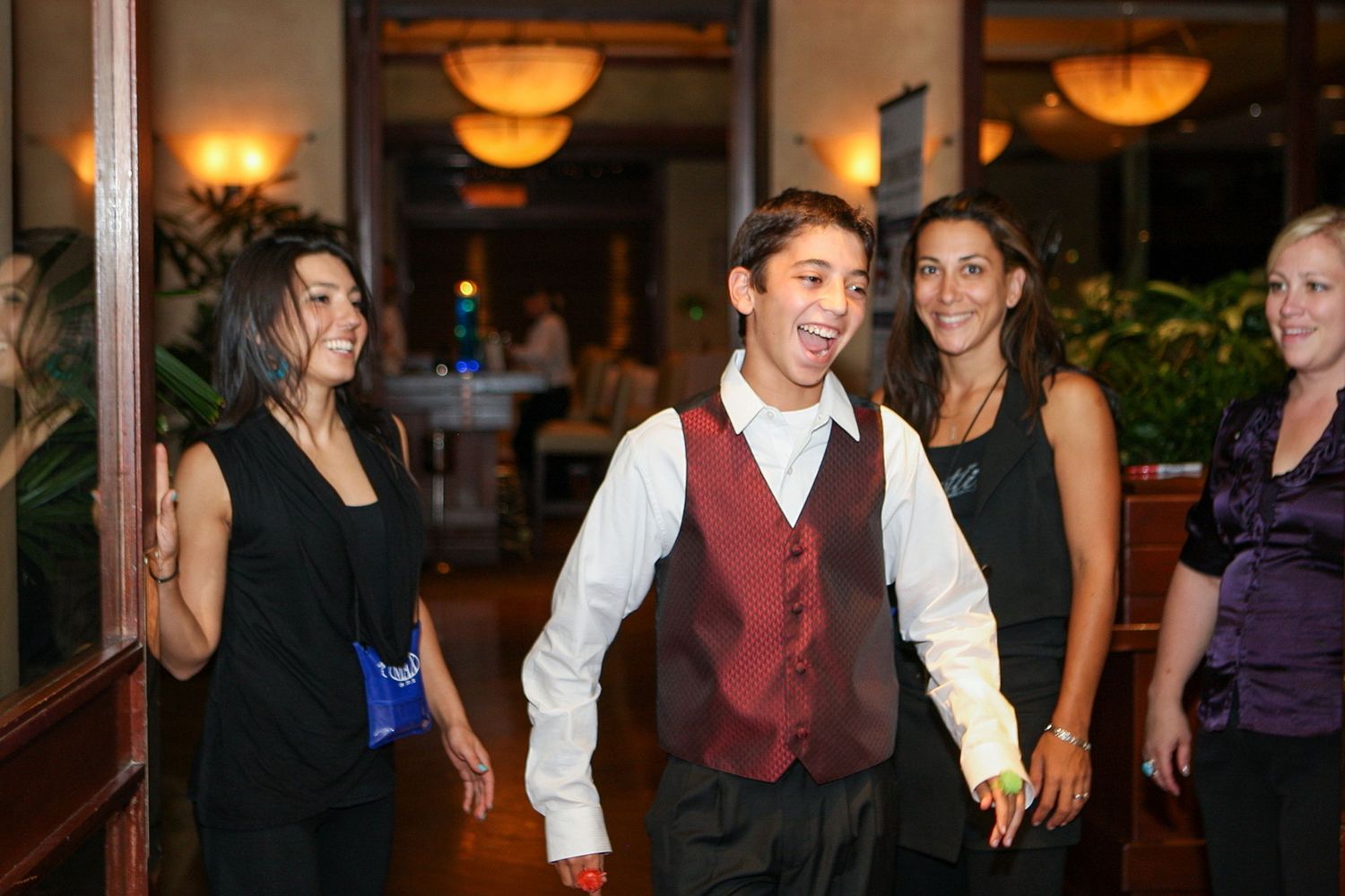 Young waiter in red vest smiles while walking with guests in an elegant restaurant setting.
