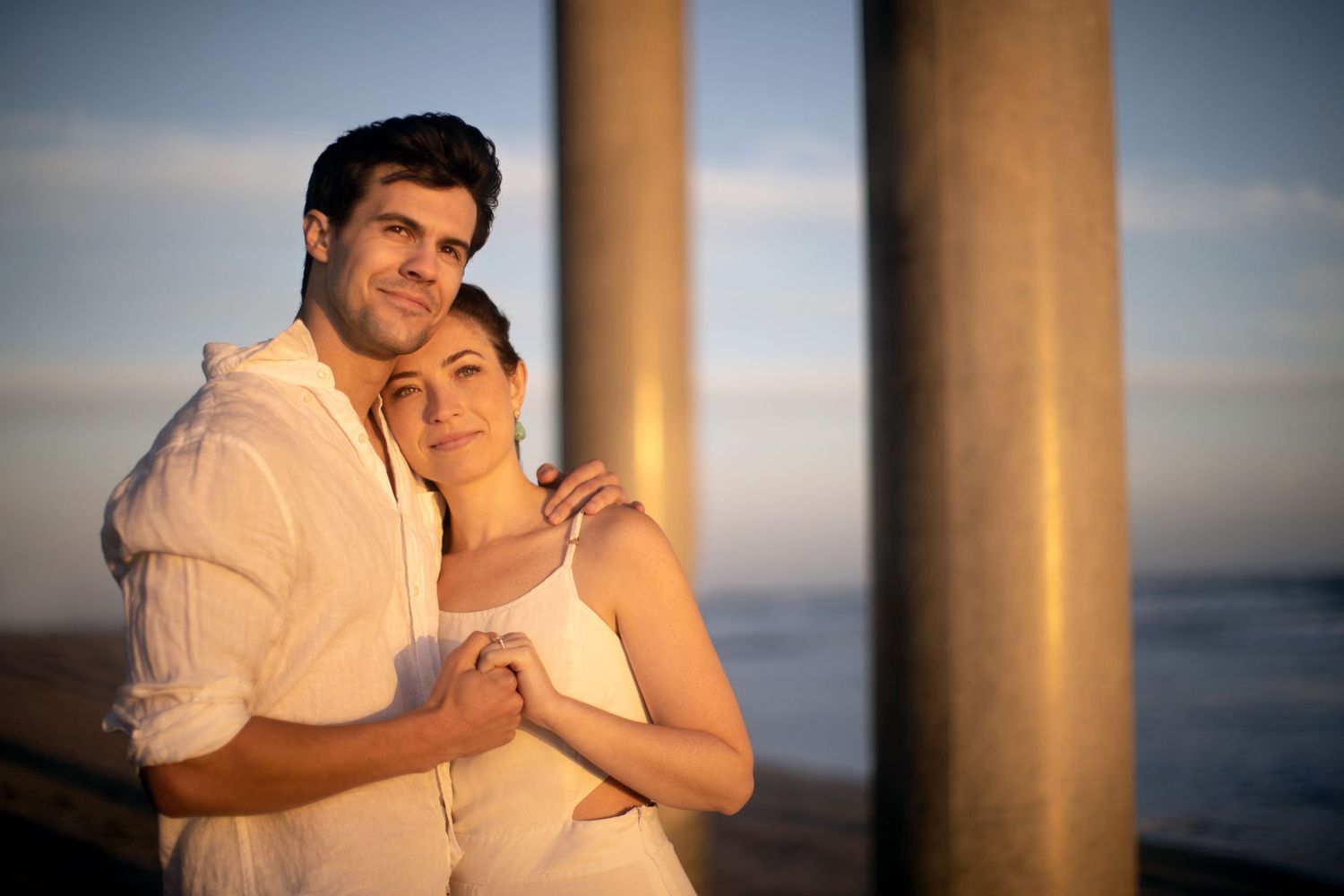 Couple in white embracing warmly near pier columns bathed in warm golden sunset light.