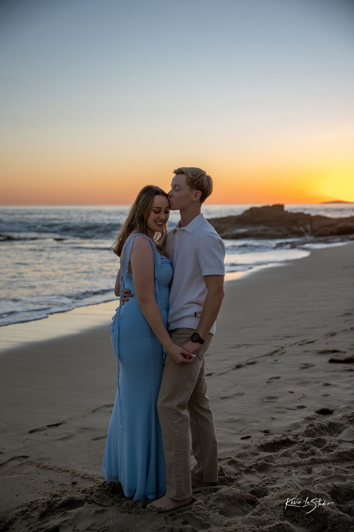 Man kissing woman's cheek on beach at sunset, woman in blue gown smiling.