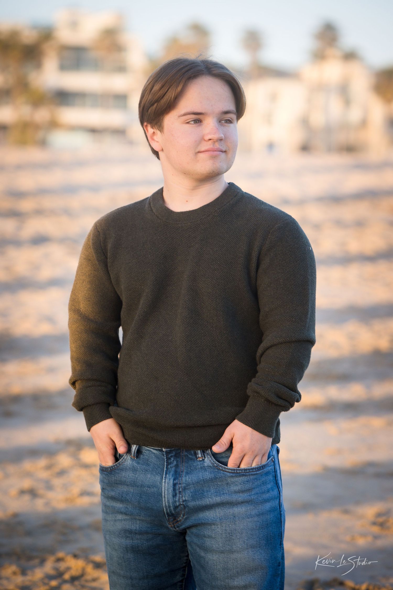 Young man in dark green sweater posing on sunny beach looking away.