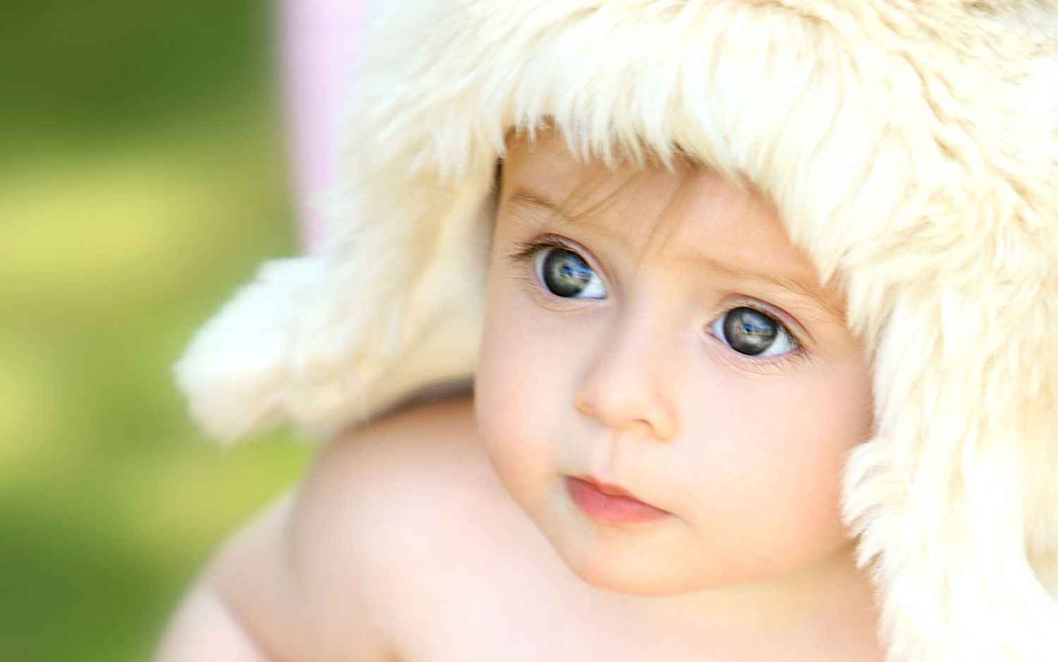 Adorable baby with big blue eyes wearing a fluffy white hat, looking curiously at the camera.