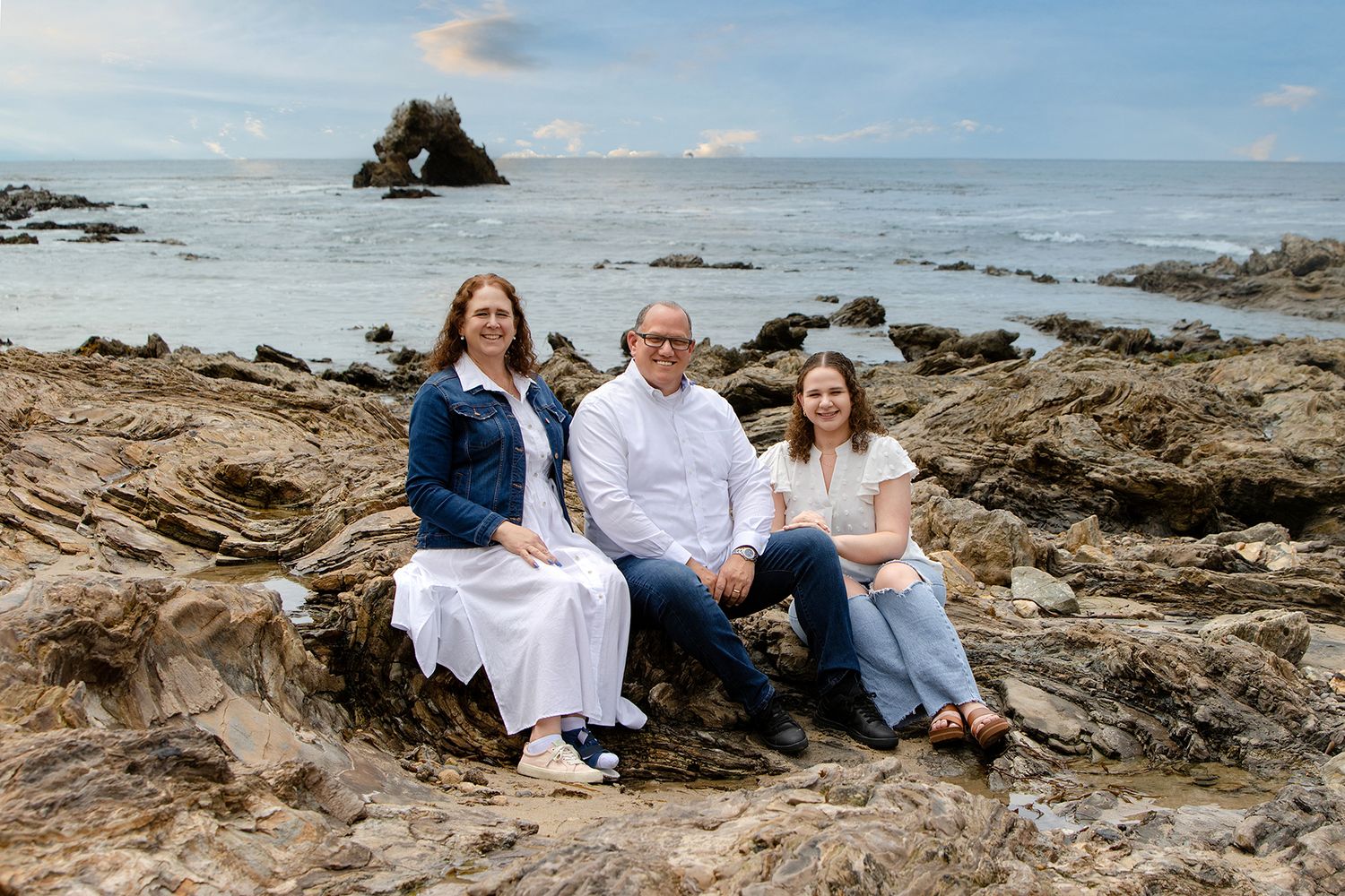 Family of three smiling on rocky coastal shore with ocean waves and rock formations in background.