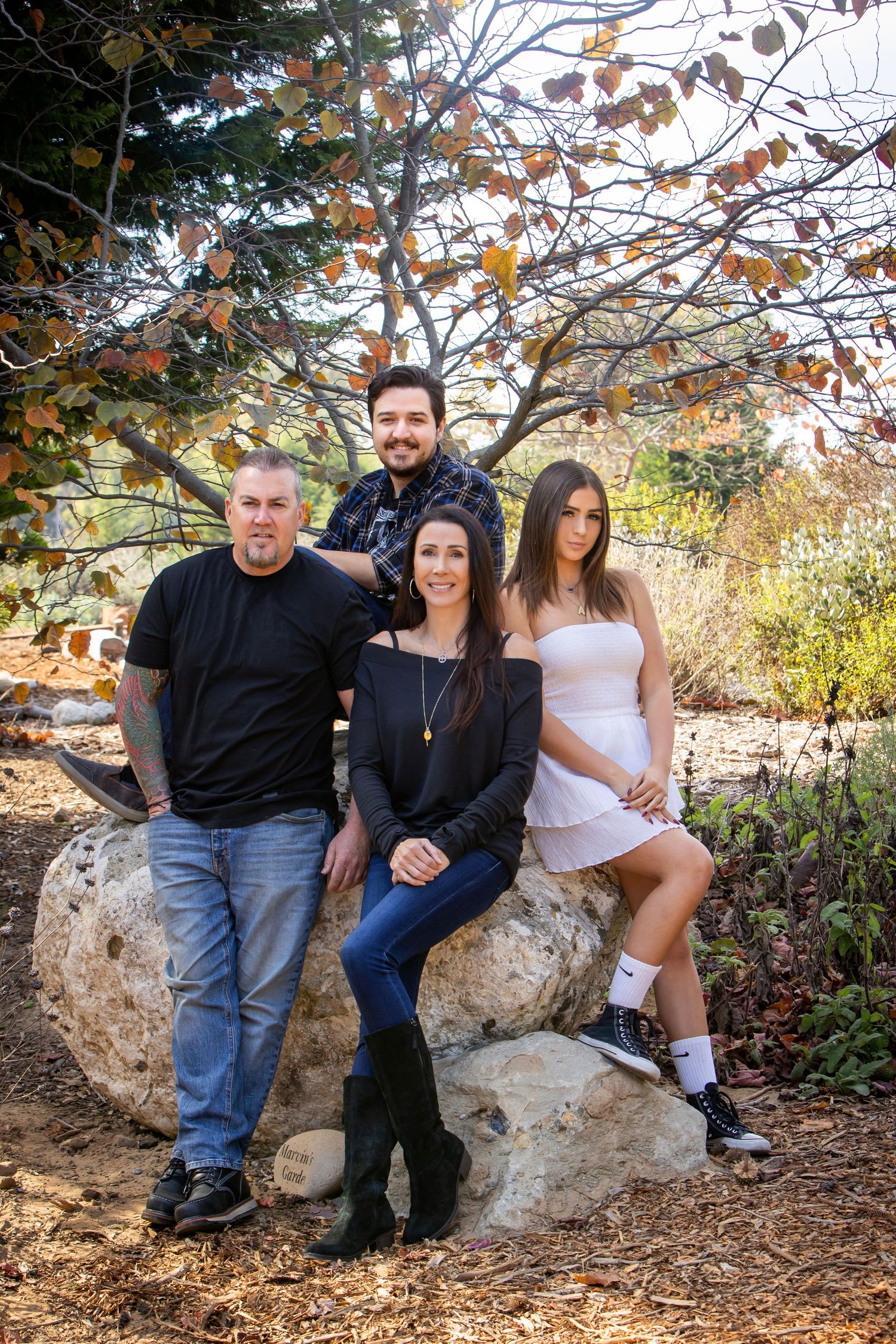 Family of four posing on a large rock outdoors beneath a tree with autumn foliage in a garden setting.