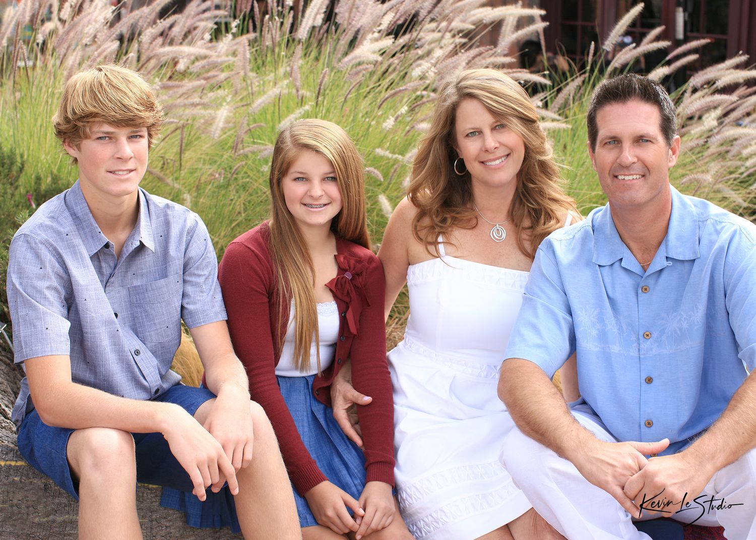 A smiling family of four poses outdoors, featuring two teenagers and their parents dressed in casual summer attire.