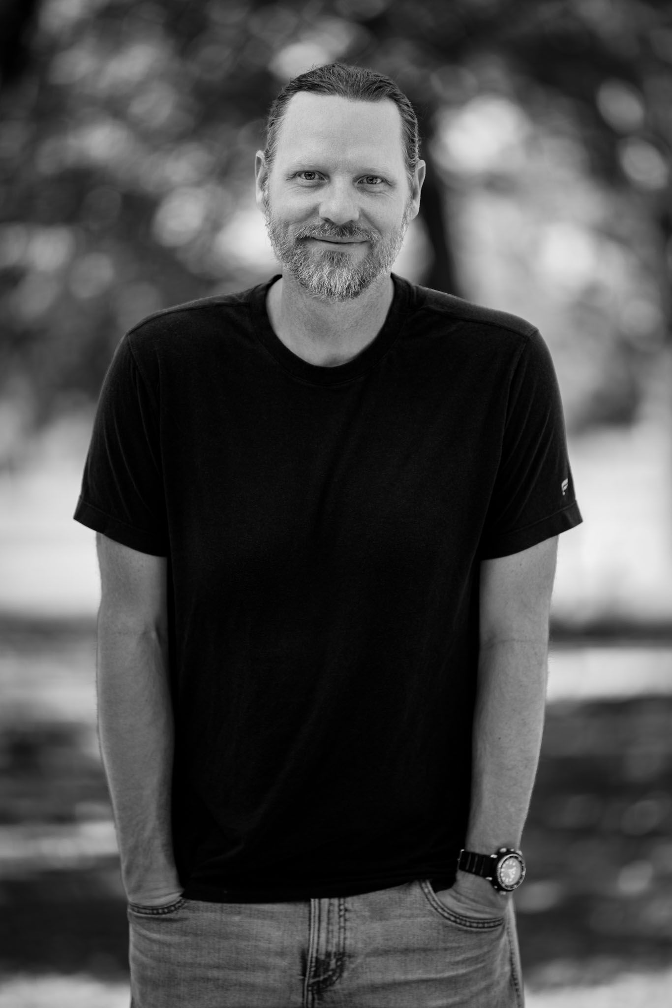 Black-and-white portrait of a bearded man smiling with hands in his pockets in a park