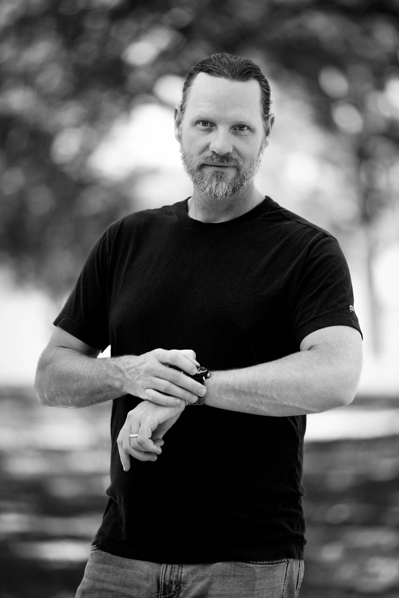 Black-and-white lifestyle portrait of a bearded man adjusting his wristwatch under trees