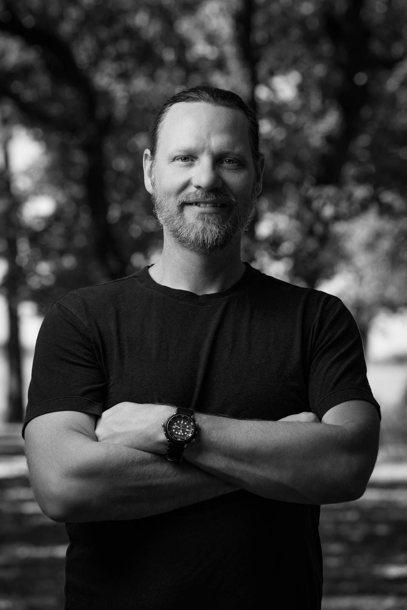 Black-and-white portrait of a smiling bearded man with arms crossed, trees behind him