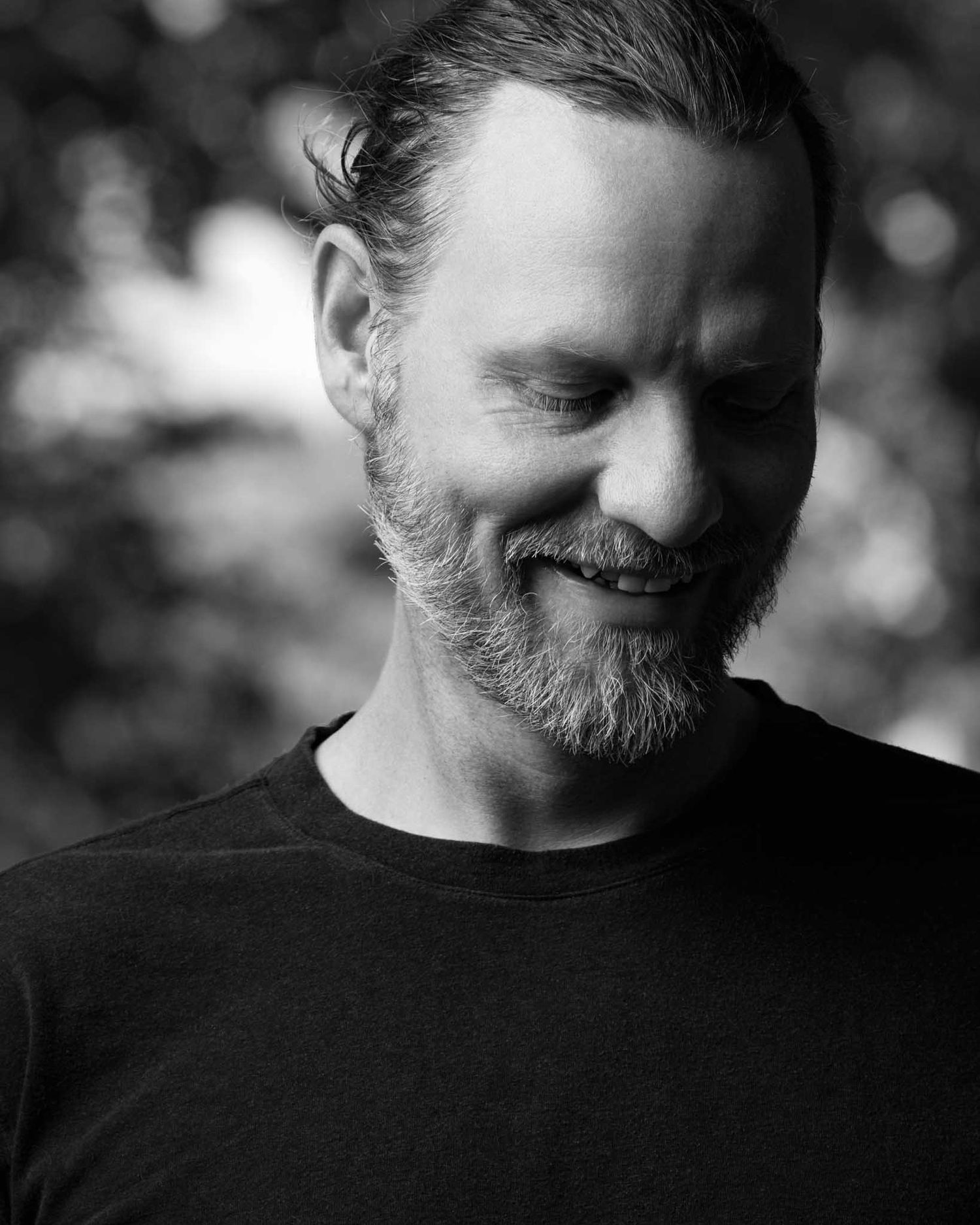 Moody black-and-white close-up of a bearded man with slicked hair looking down and smiling, dark foliage