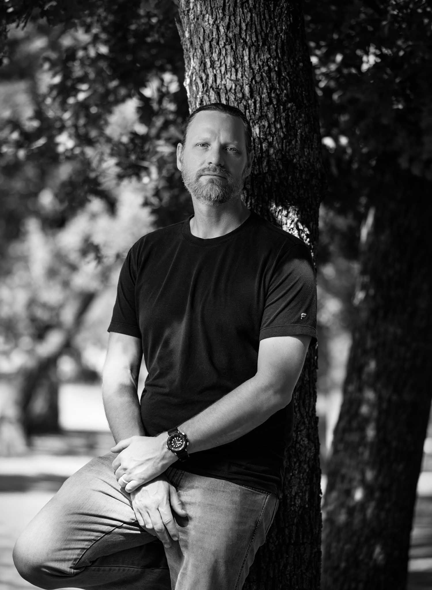 Black-and-white portrait of a bearded man in a black tee leaning against a tree in a shady park