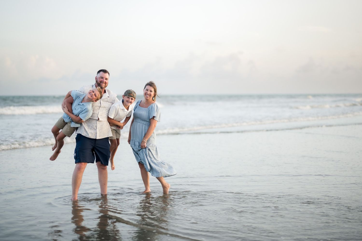 Family of four laughing and playing on a sandy beach with ocean waves, wearing light blue and gray outfits.