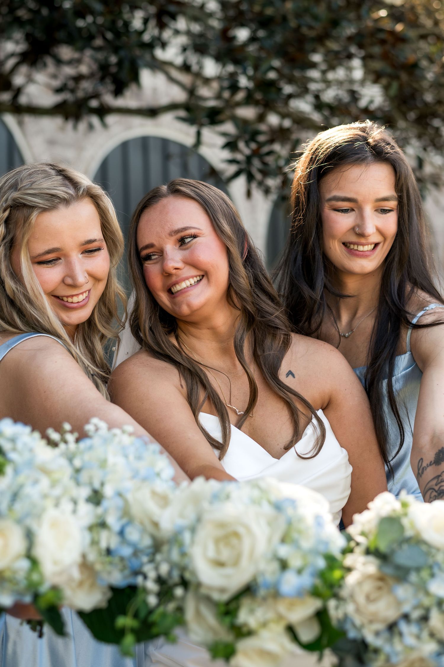 Dunes West Golf and River Club. Smiling bride with two bridesmaids in light blue dresses holding white and blue floral bouquets outdoors.
