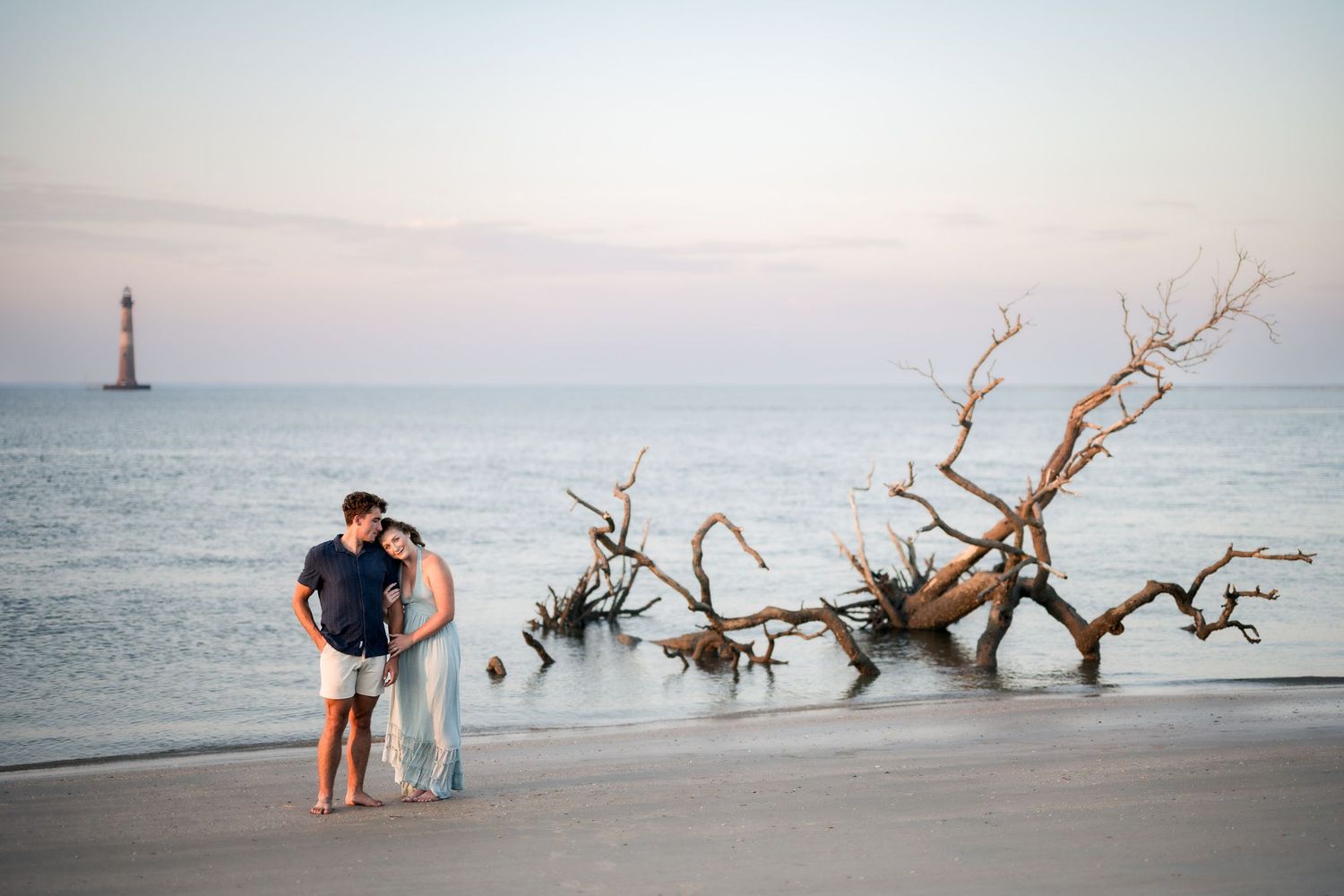 Couple embraces on a beach at sunset near driftwood with a lighthouse visible in the distance.