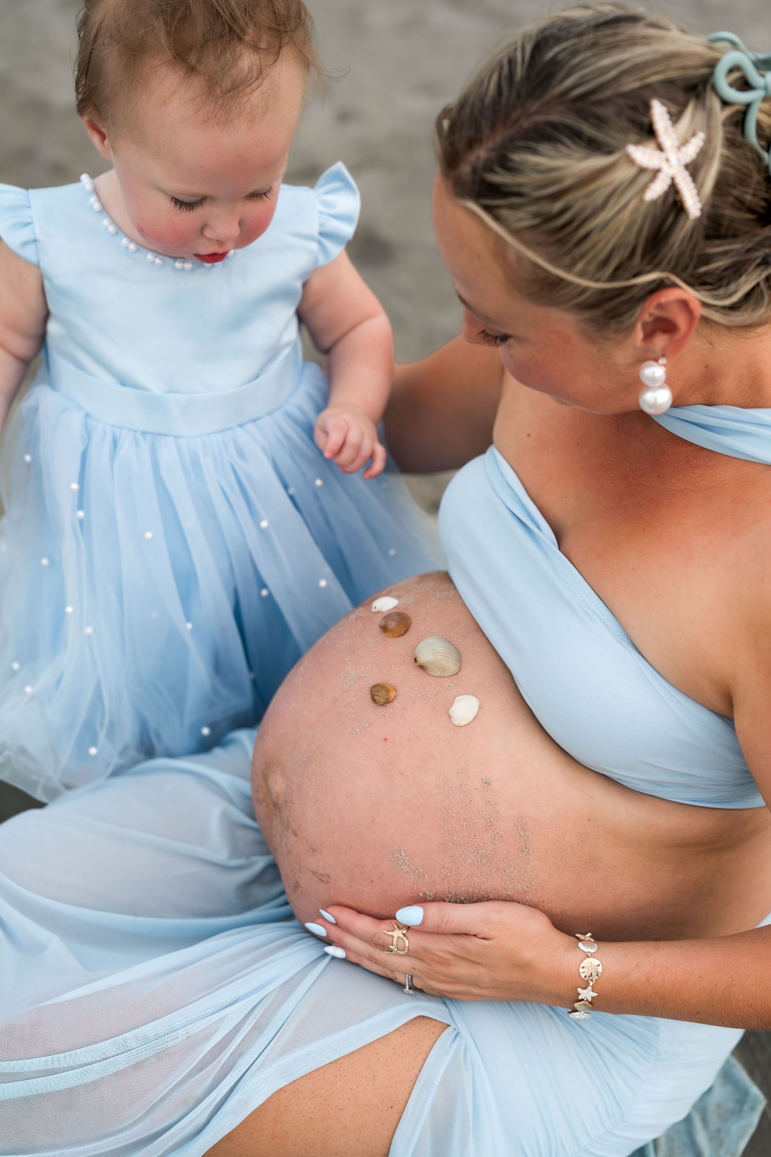 Pregnant woman in blue dress with toddler girl placing seashells on her baby bump during a beach maternity photoshoot.