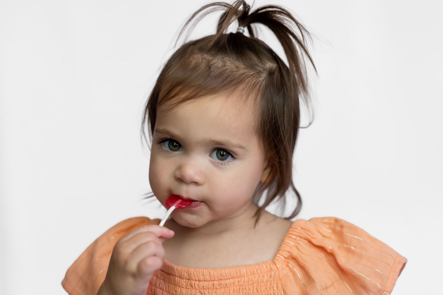 Toddler girl with ponytail in orange dress eating a red lollipop against white background.