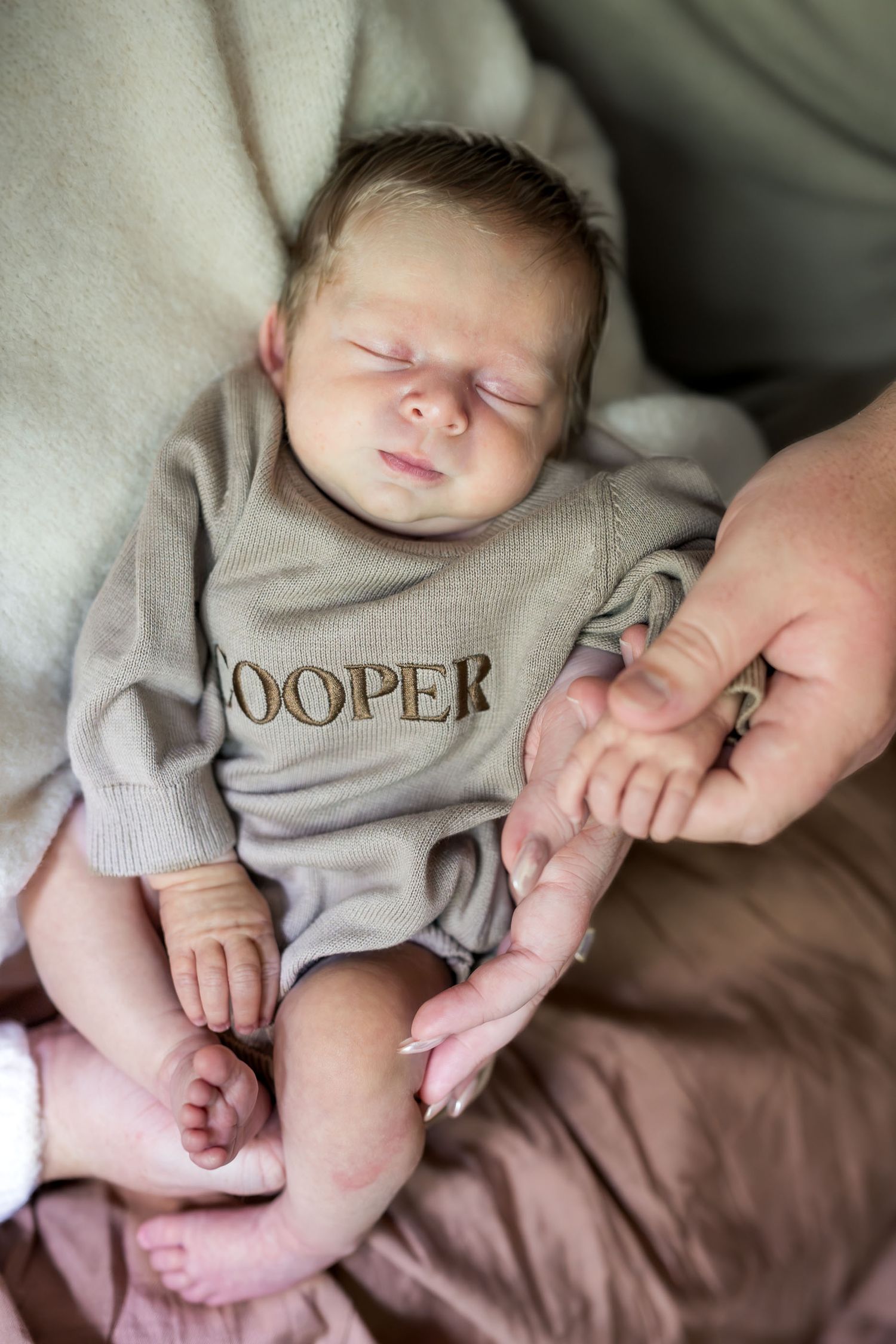 Sleeping newborn baby named Cooper in a gray personalized onesie, gently holding an adult's finger.