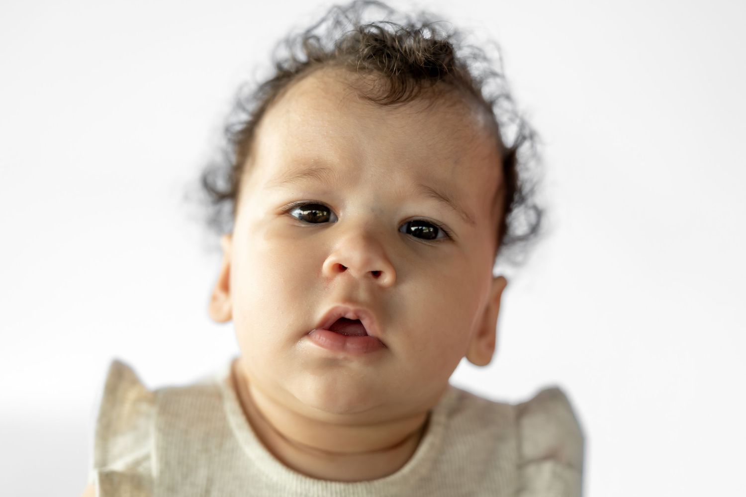 Close-up portrait of an adorable baby with curly dark hair and chubby cheeks wearing a gold ruffled outfit.