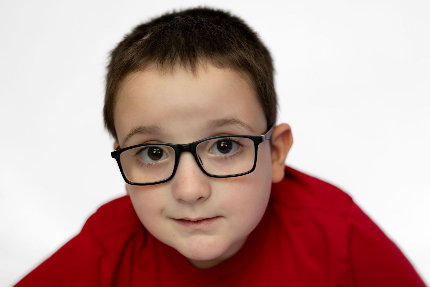 Young boy wearing black rectangular glasses and a red shirt poses for a portrait against a white background.