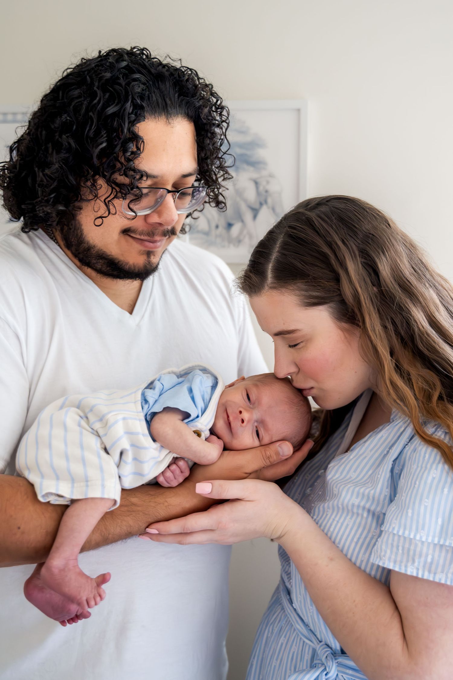 Young parents lovingly hold their newborn baby, with mother kissing infant's head in a bright home setting.