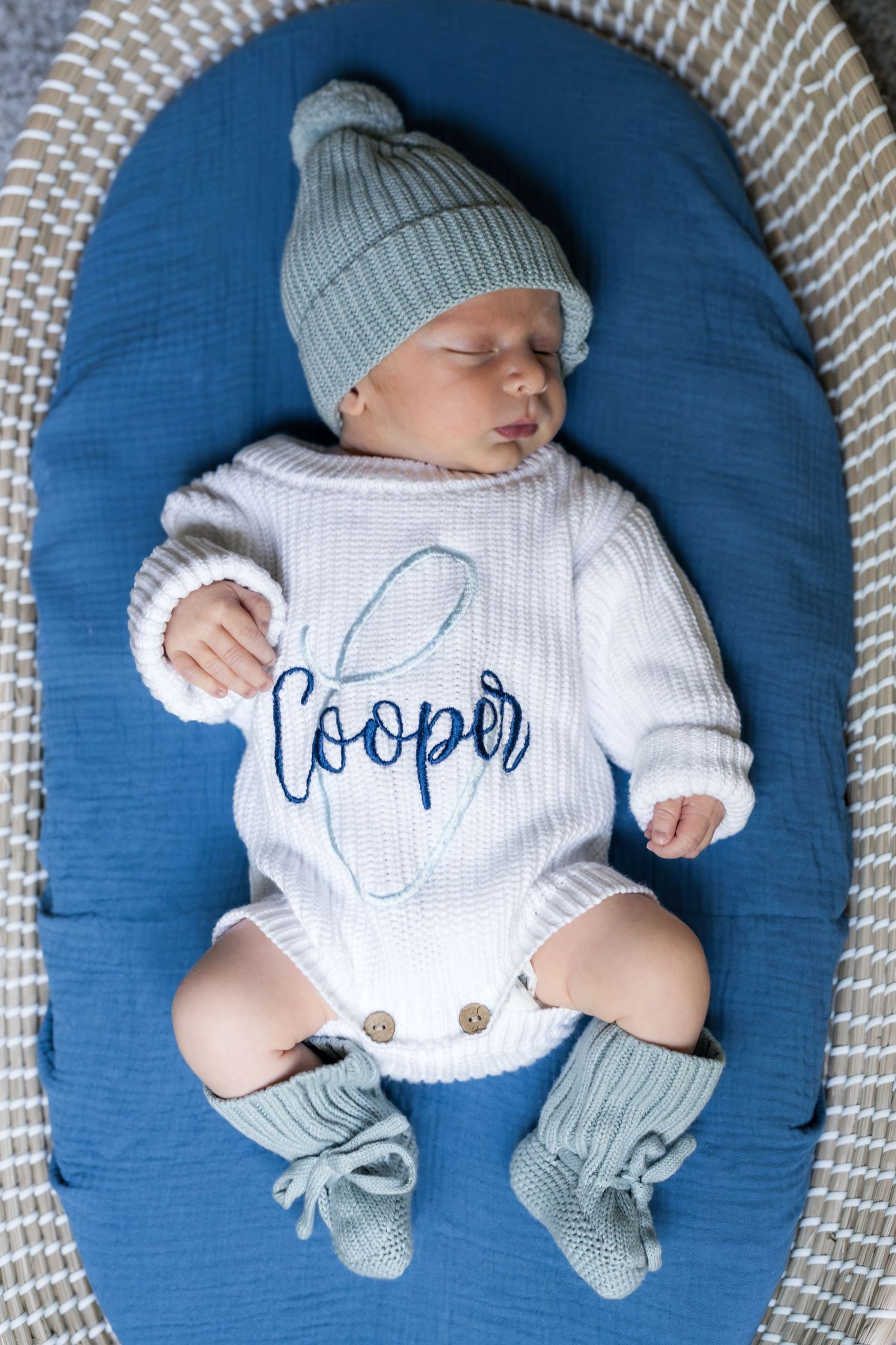 Newborn baby named Cooper sleeping in blue nest, wearing white knit romper, sage green hat and booties.