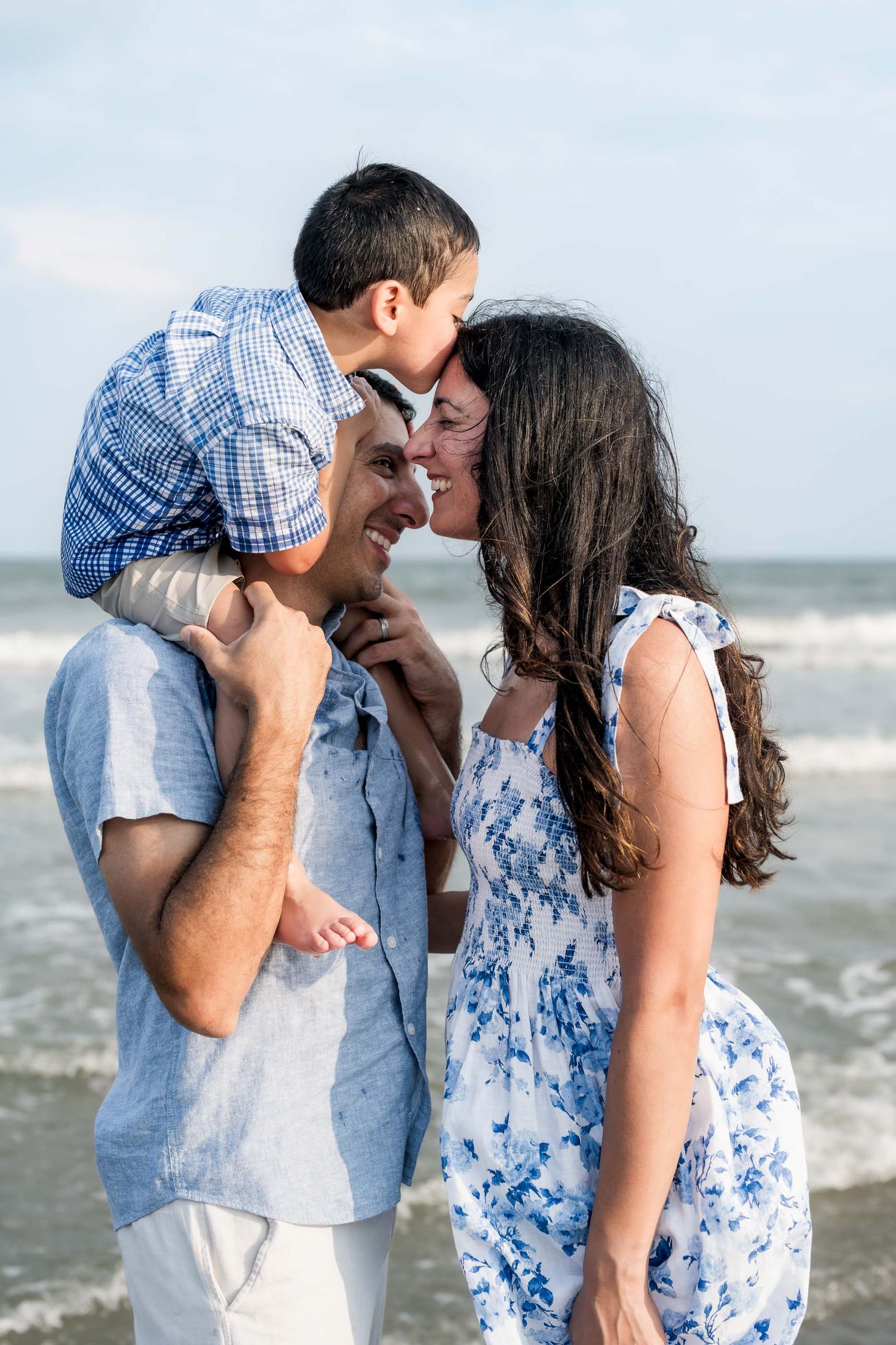 A happy family at the beach, with a boy on his father's shoulders as the mother leans in for a kiss.