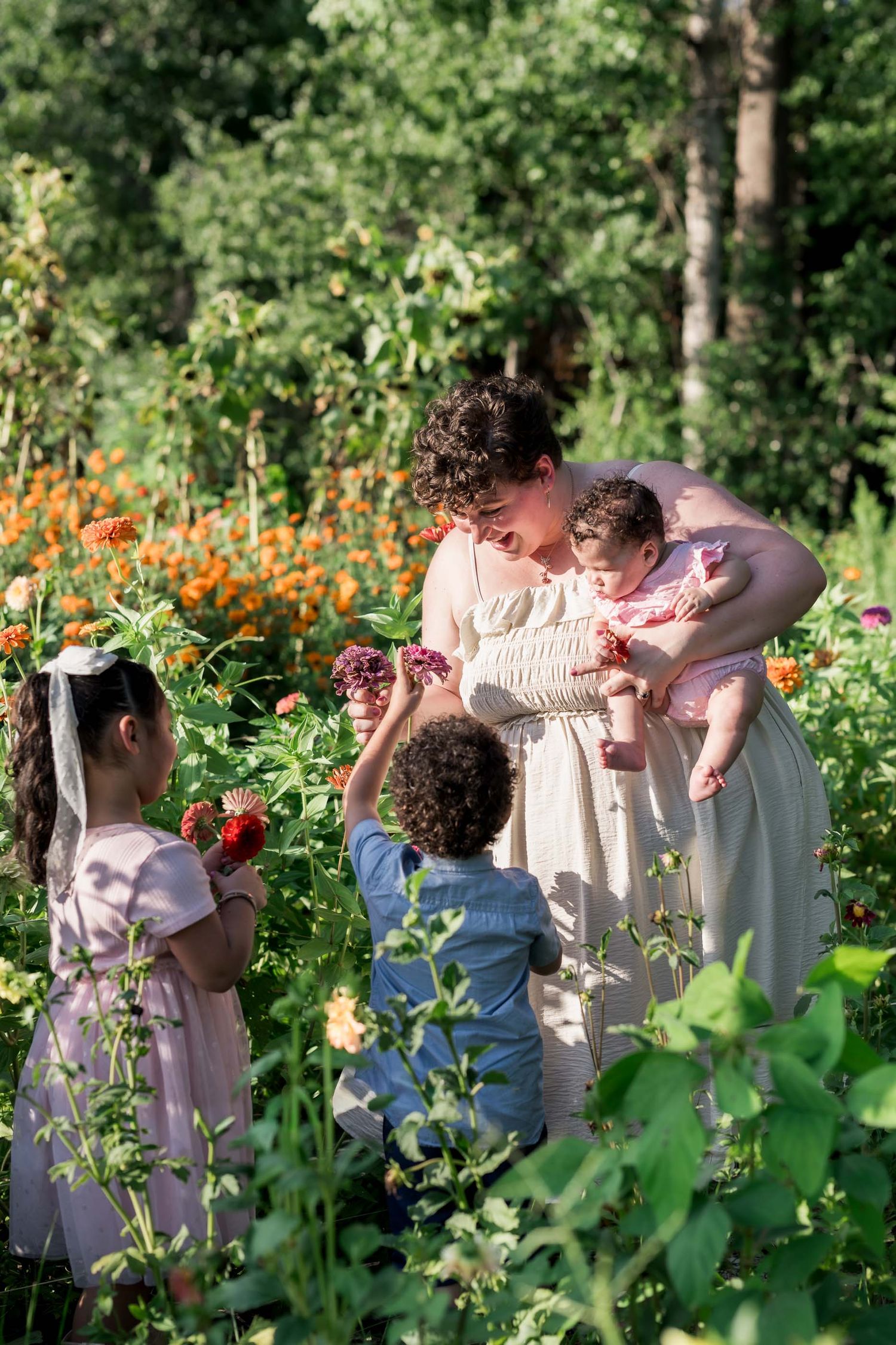 A mother in a white dress holds a baby while two young children offer flowers in a lush garden.