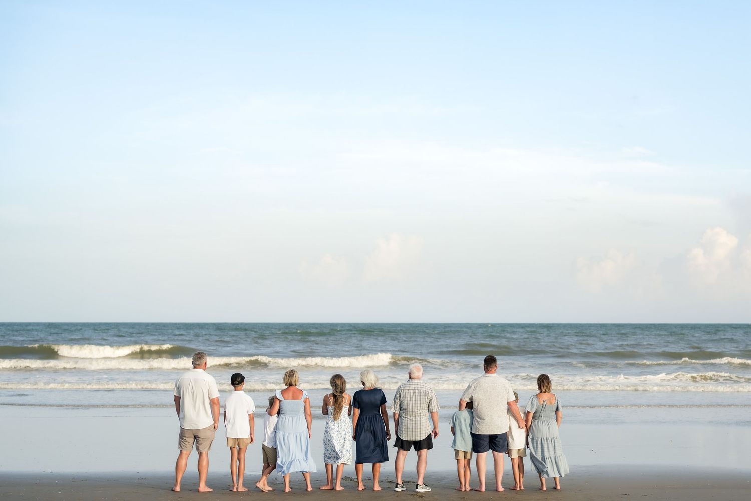 Multi-generational family standing together on a sandy beach, facing the ocean waves under a clear blue sky.