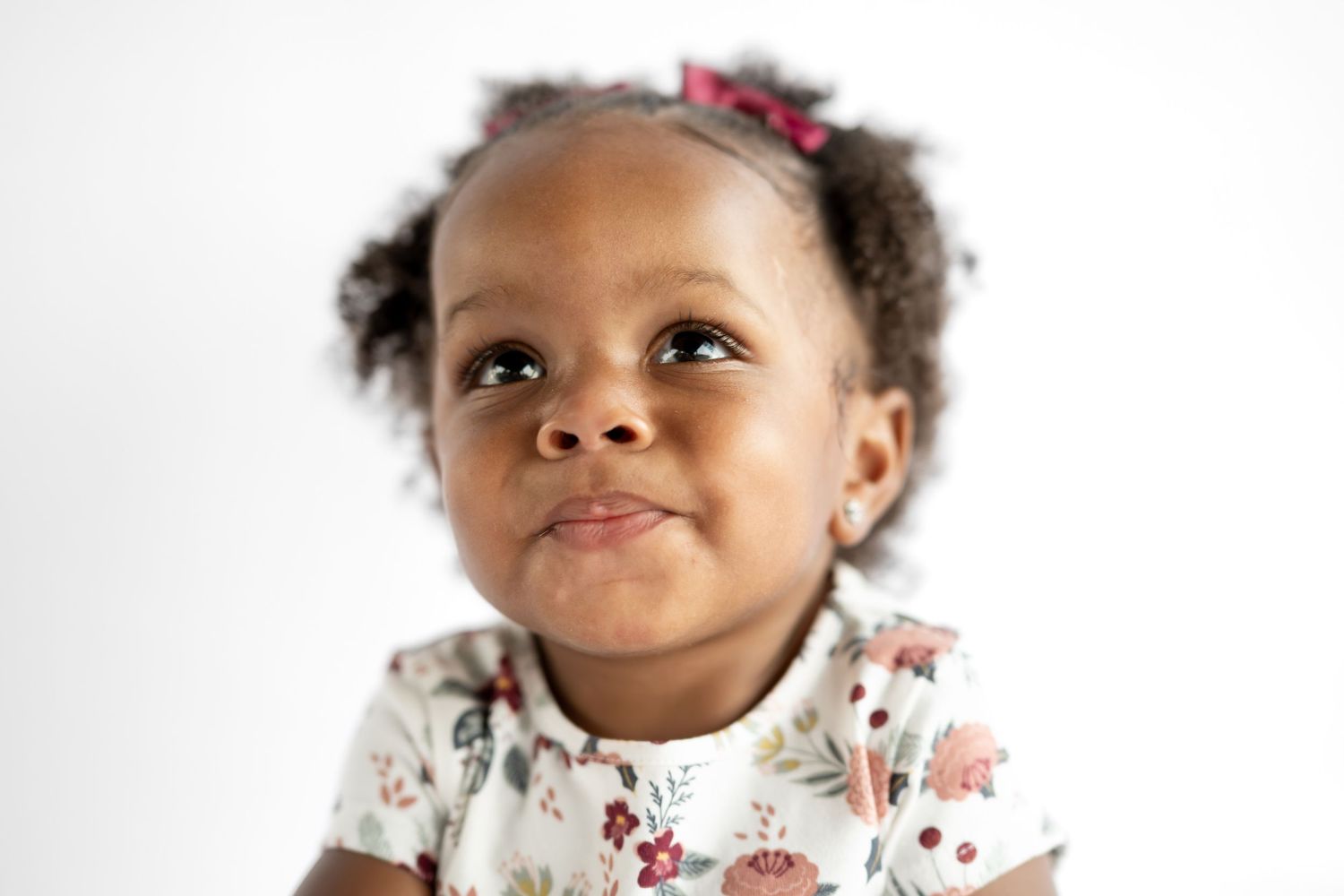 Adorable Black toddler girl with curly hair and pink bow smiling upward, wearing floral dress on white background.