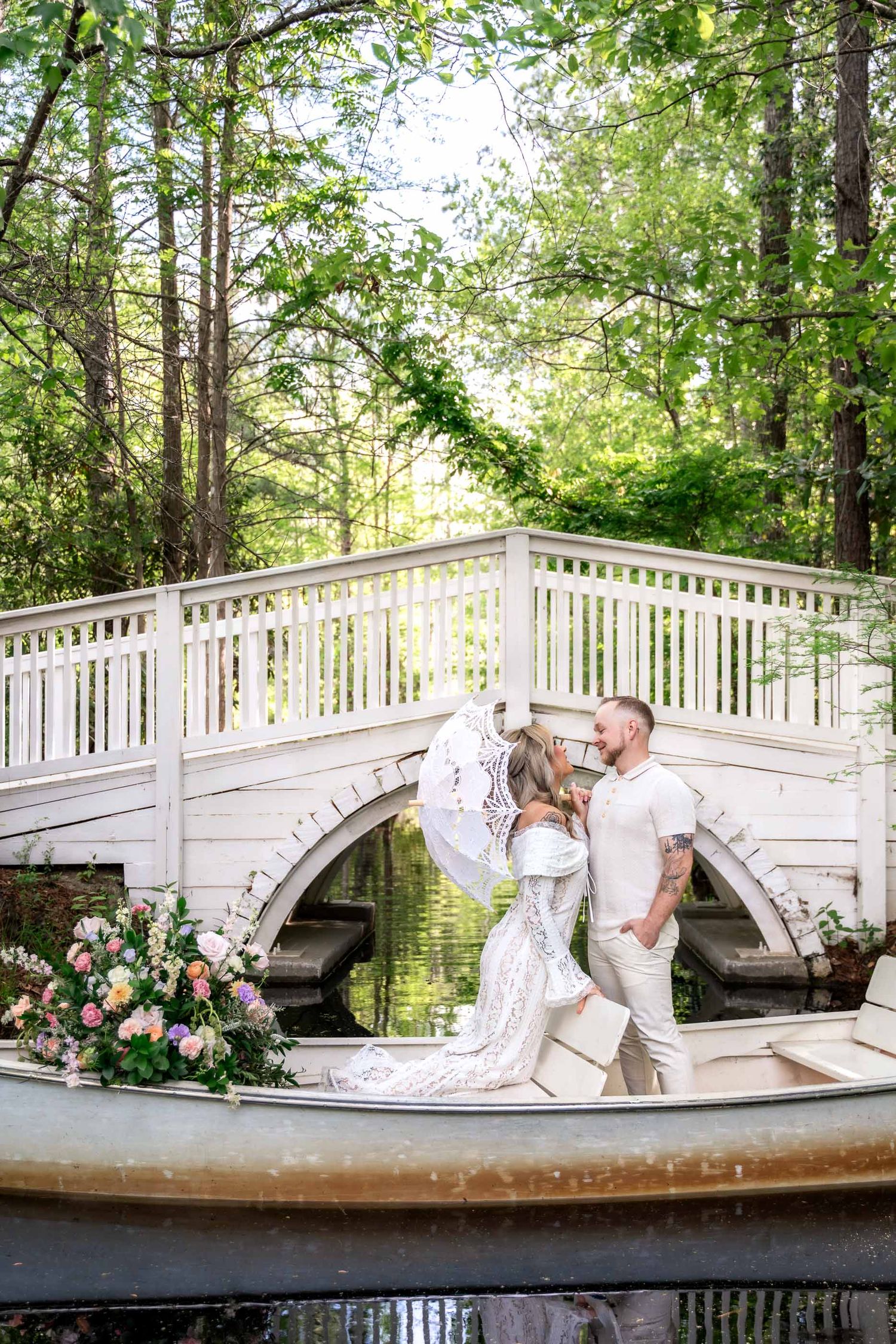 Couple sharing a romantic kiss in a white rowboat near a charming white bridge surrounded by lush green trees.