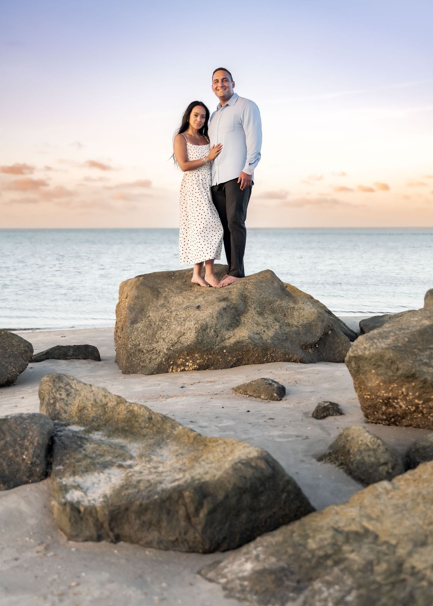 Folly Beach. Romantic couple standing on rocks at beach during sunset, woman in white dress, man in light blue shirt.