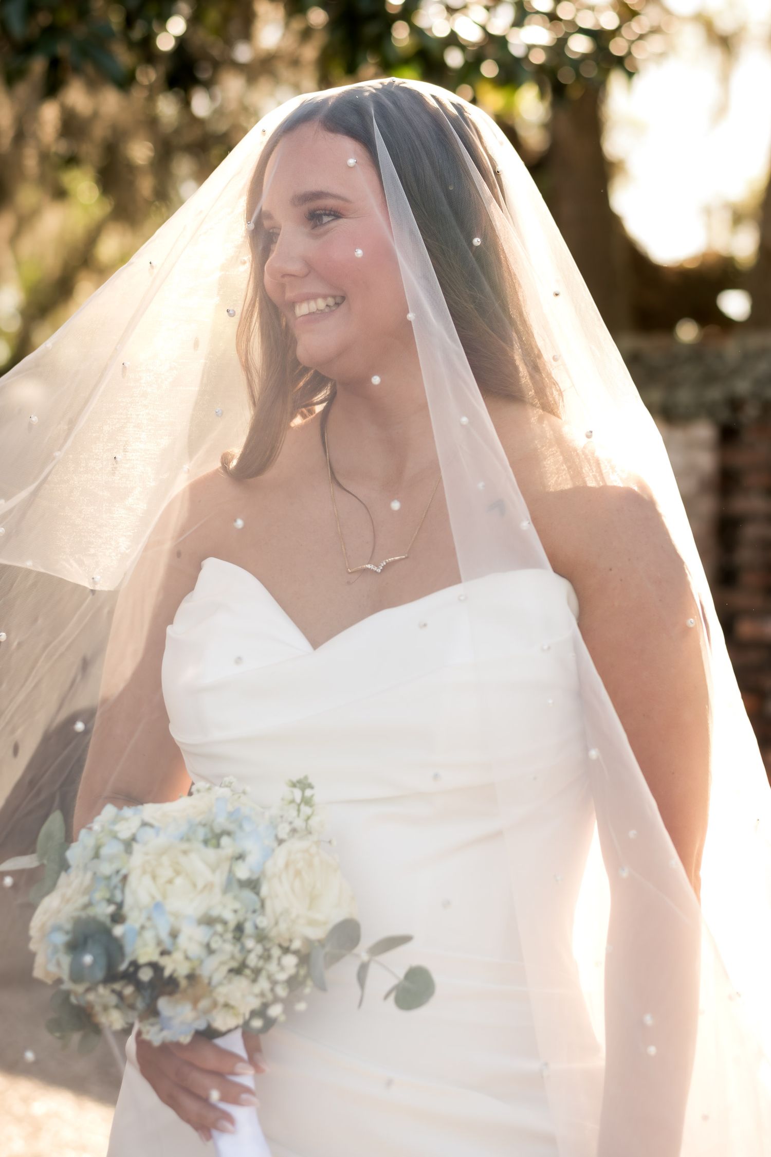 Dunes West Golf and River Club. Radiant bride in strapless white dress and pearl-dotted veil holding white bouquet in golden sunset light.