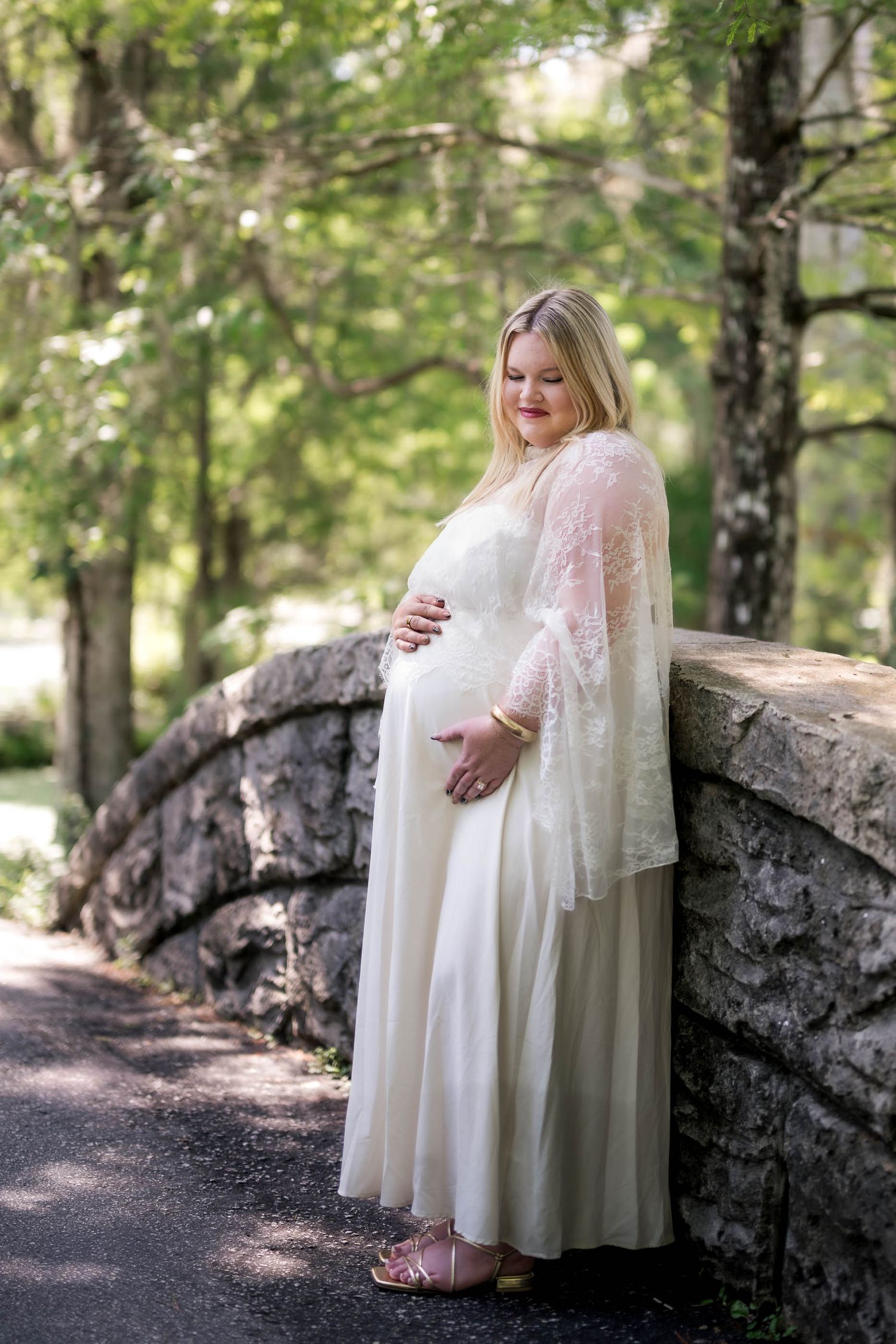 Pregnant woman in white lace gown cradling baby bump beside stone bridge in lush green park setting.