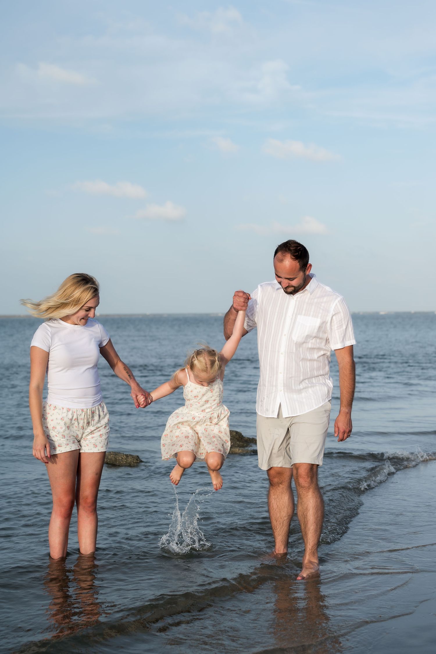 Family of three enjoying beach, parents swinging toddler over ocean waves while walking barefoot on sandy shore.