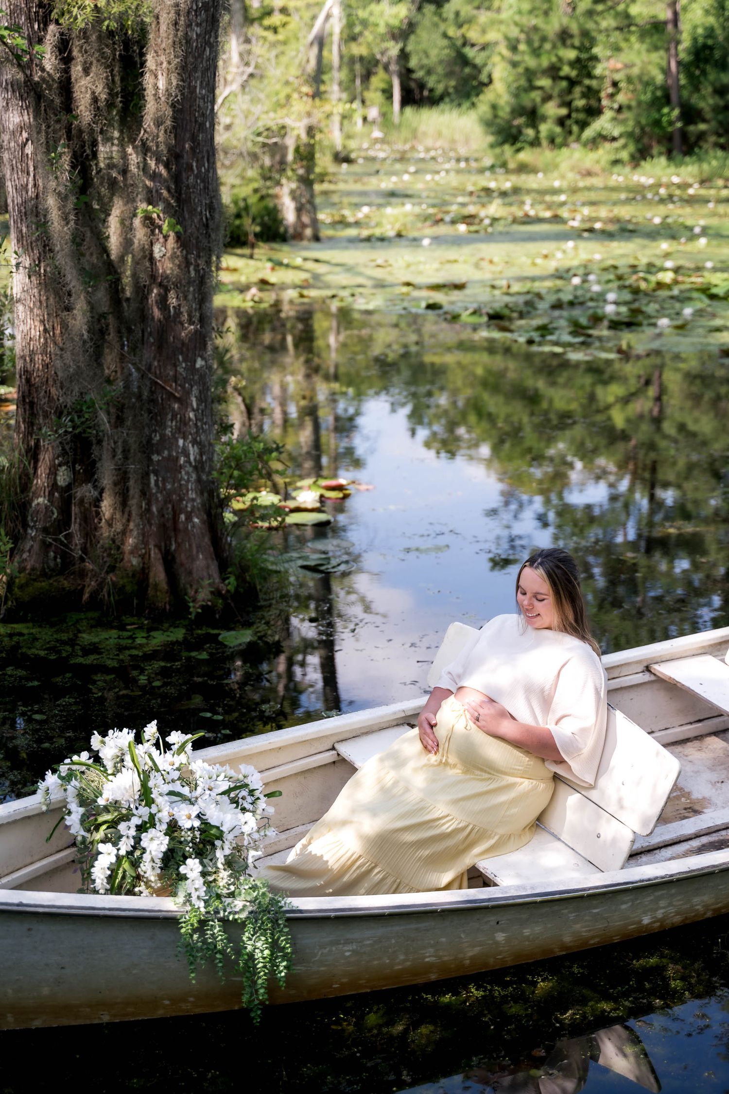 Woman in yellow dress reclines in a wooden boat surrounded by lily pads and white flowers on a serene pond.