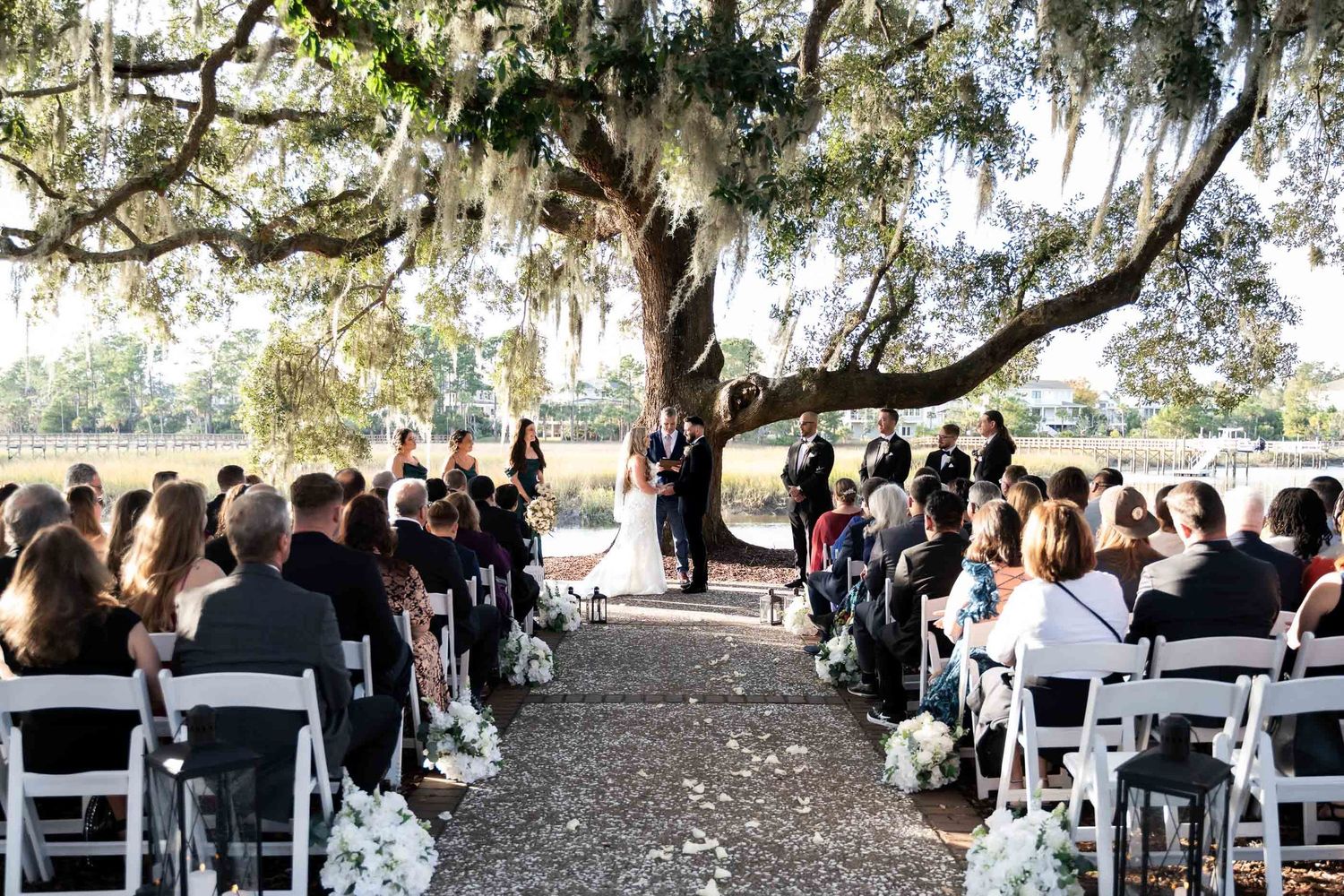 Dunes West Golf and River Club. Outdoor wedding ceremony under a sprawling Spanish moss oak tree with white chairs and floral aisle decor.